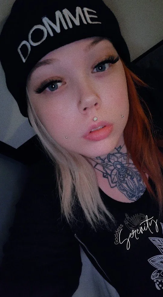 A young woman with tattoos on her neck, multiple facial piercings, and light blonde and red hair, wearing a black beanie with 'DOMME' written on it and a black shirt with a design and script visible.
