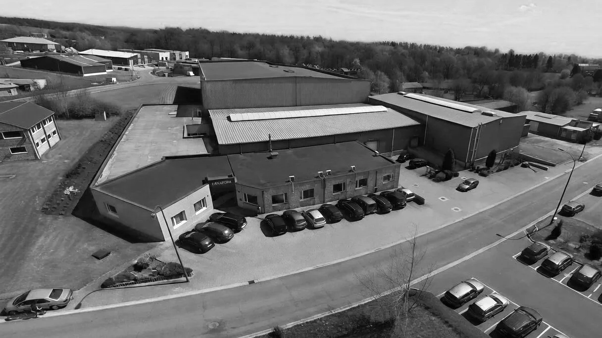 Aerial view of a commercial building with cars parked in front, surrounded by a parking lot, other buildings, and trees in the background.