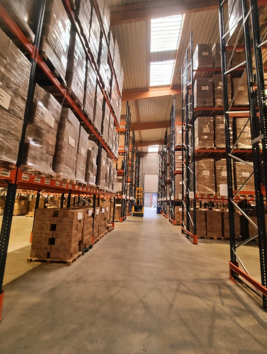 Warehouse aisle with tall shelving units filled with stacked boxes, some wrapped in plastic, and a forklift at the back near an open door.
