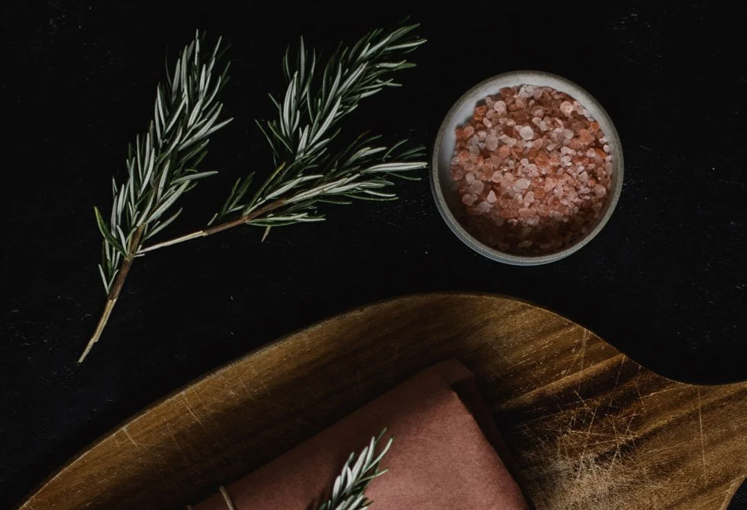 Klip på rosmarin, en skål med pink Himalayasalt og en træbakke med brunt papir på en sort overflade.