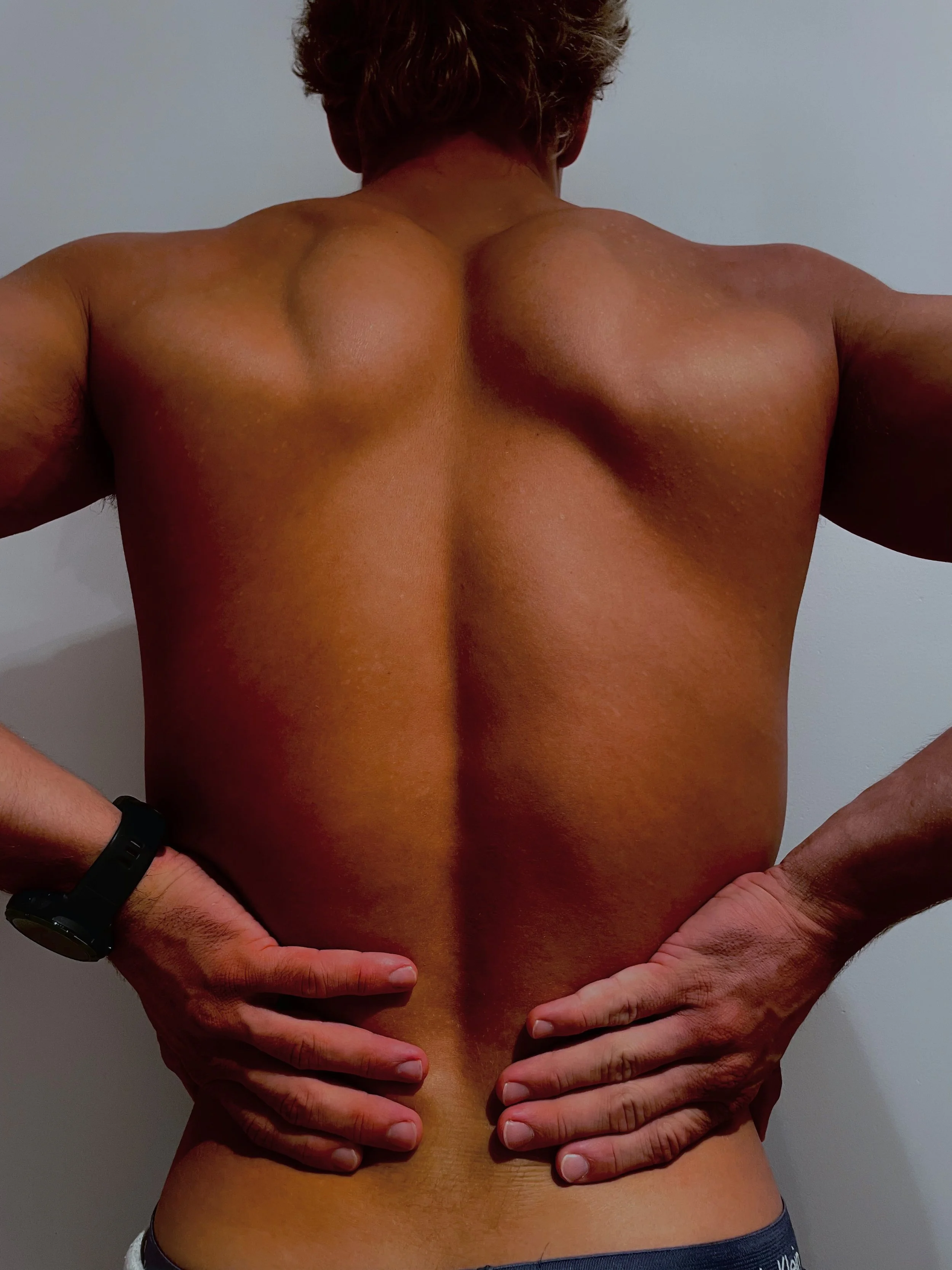 A person's bare back with hands placed on hips, showing muscular shoulders and upper back, against a plain wall background.