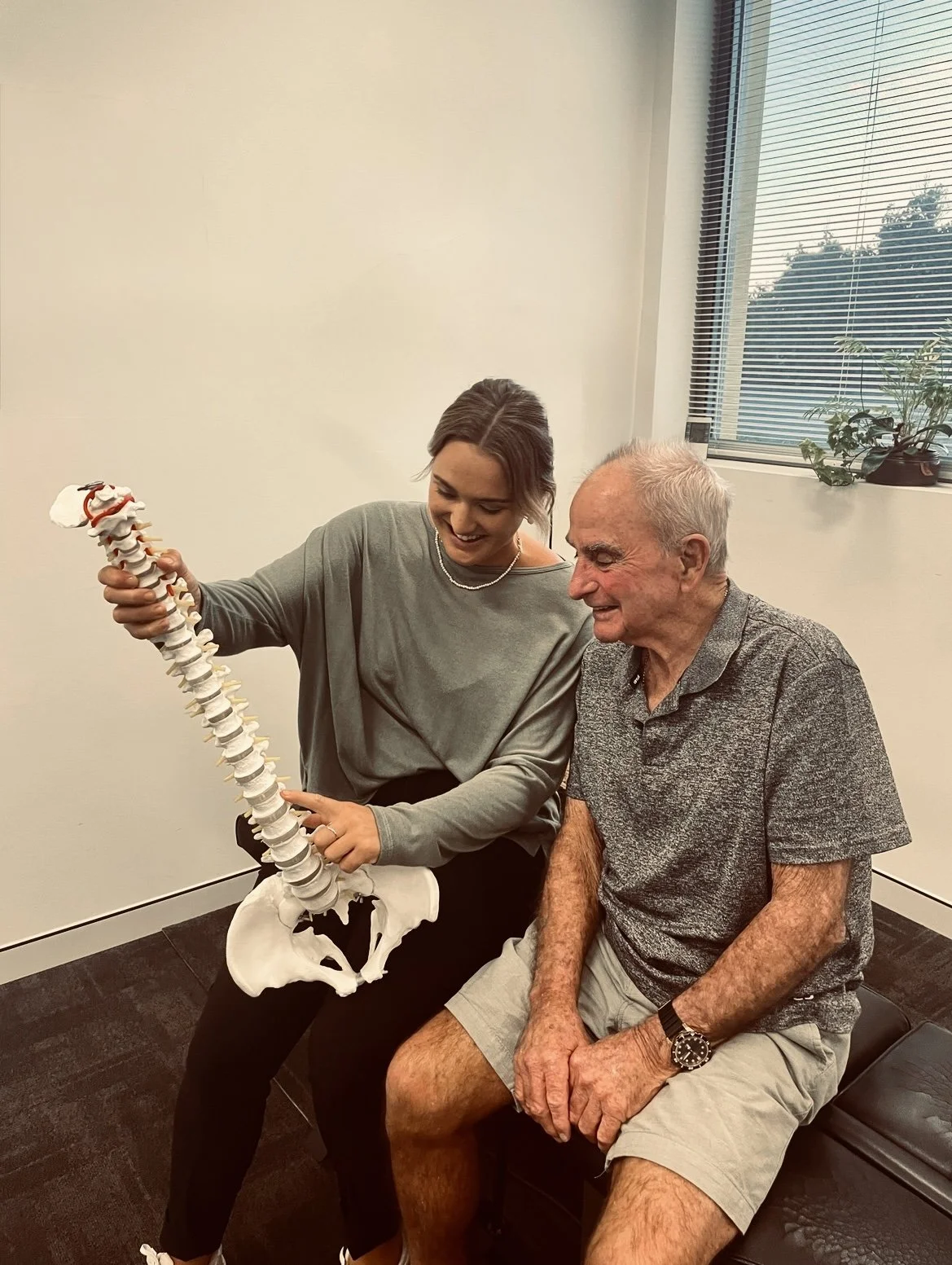 A young woman showing an elderly man a model of a human spine, both smiling in a doctor’s office.