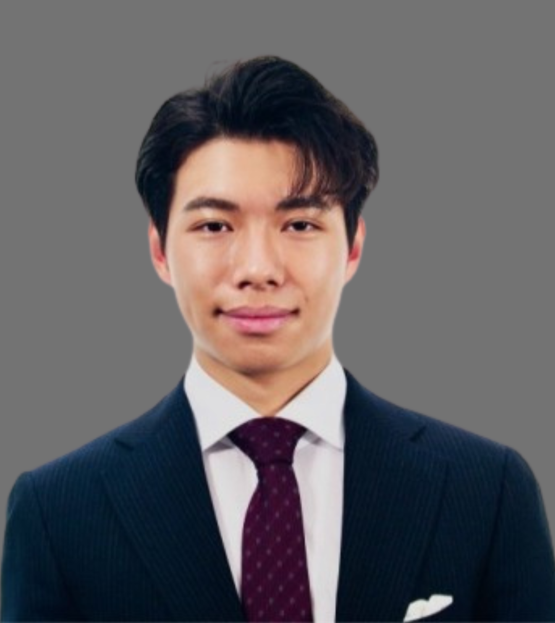 A young man in a dark suit, white shirt, and maroon polka dot tie, posing against a gray background.