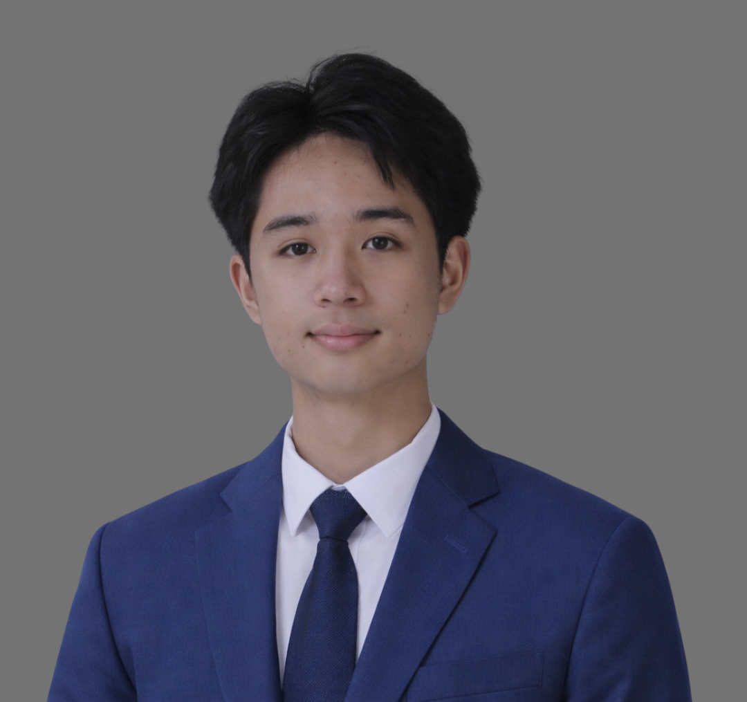 A young man with dark hair wearing a navy blue suit, white shirt, and dark tie, standing against a gray background.