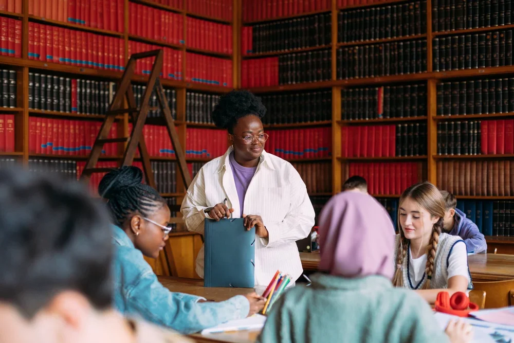 A teacher standing in a library classroom with students sitting at a table, surrounded by bookshelves filled with colorful books.