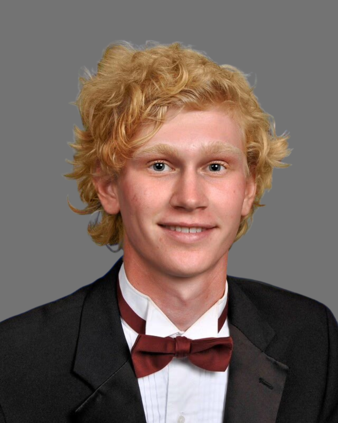Portrait of a young man with blonde, curly hair wearing a black tuxedo with a white shirt and a maroon bow tie, smiling against a plain gray background.