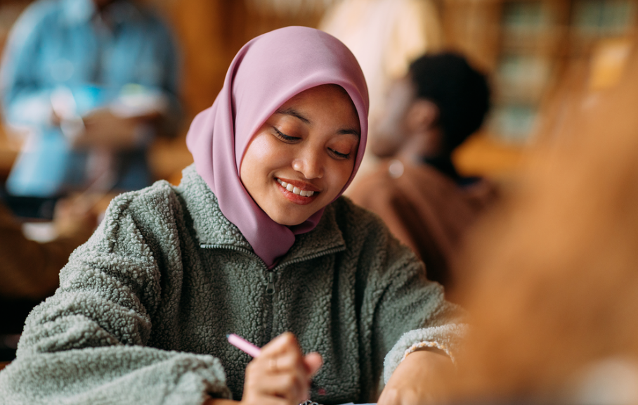 Young woman in a pink hijab and gray fleece jacket smiling while writing with a pink pen in a cozy, busy library or cafe setting.