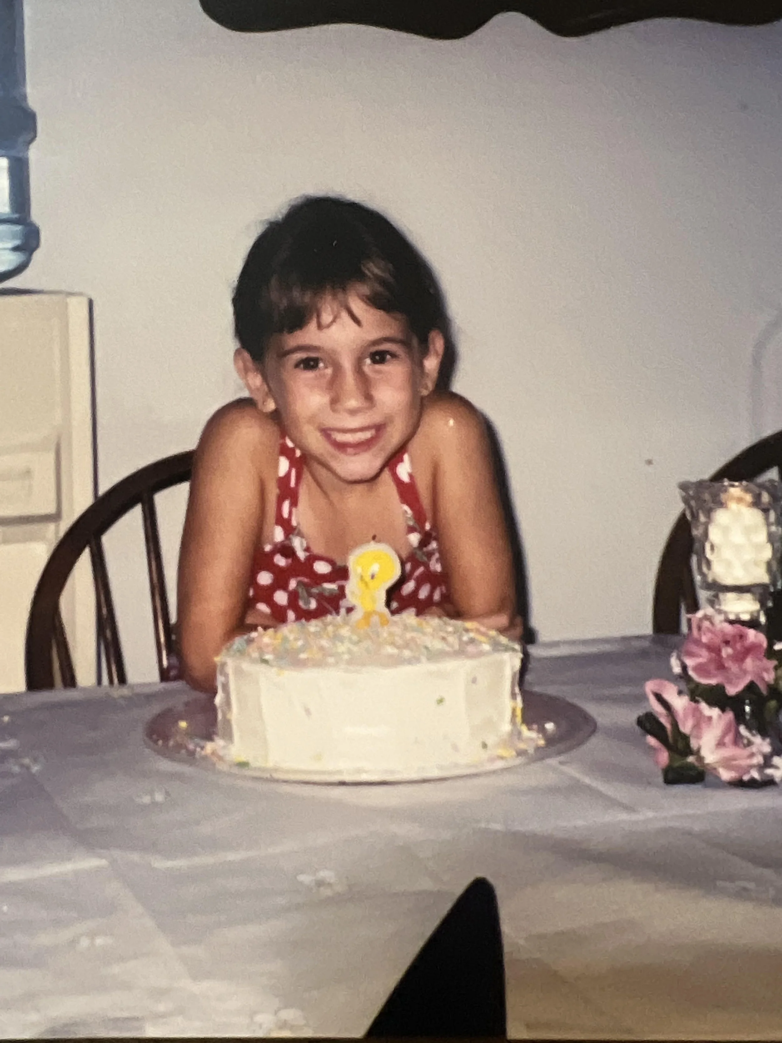A young girl with short dark hair wearing a red and white polka-dot dress is sitting at a table with a birthday cake in front of her. The cake has white icing and colorful sprinkles, and a candle shaped like the number 9. The girl is smiling.
