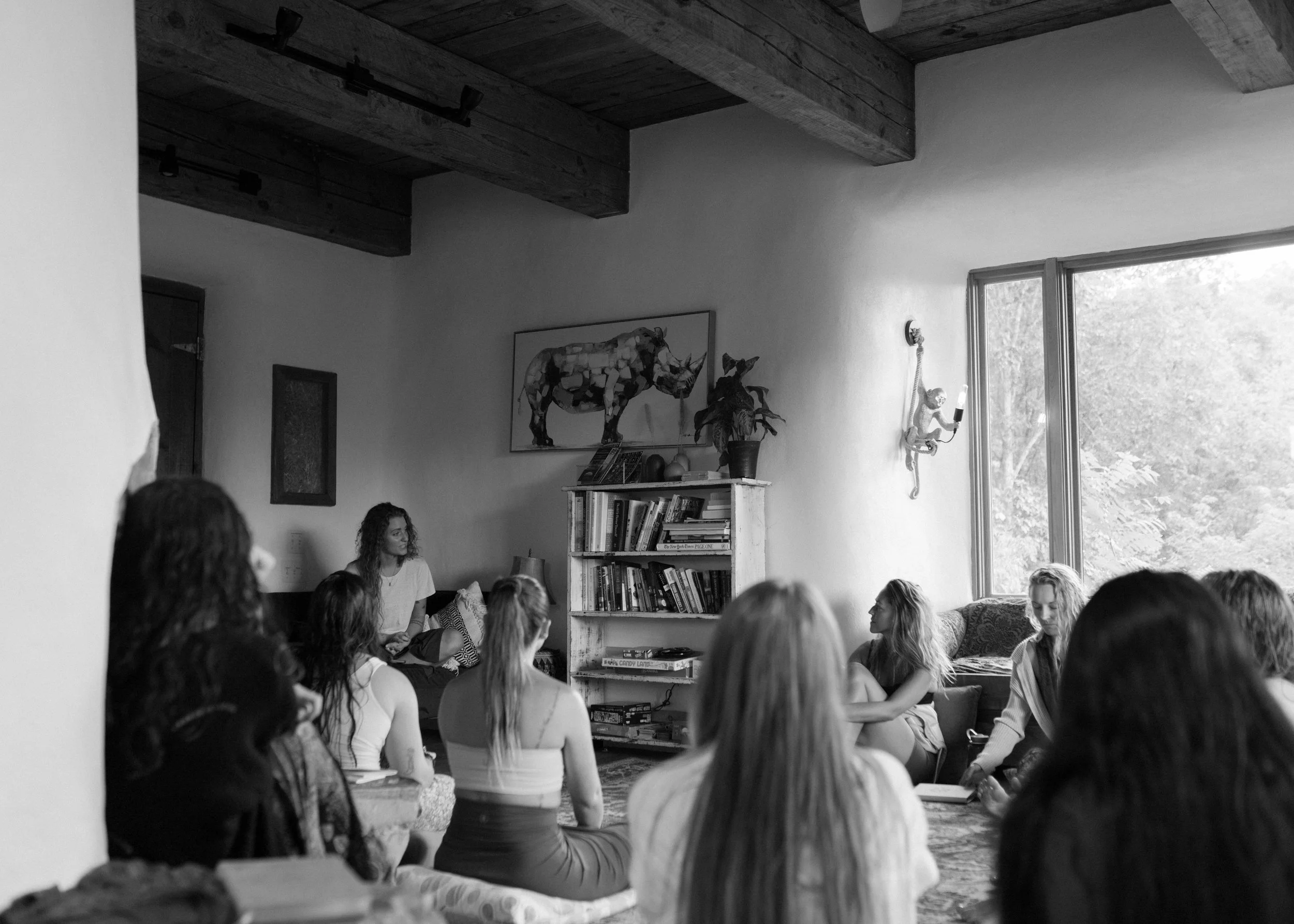 A black and white photo of a group of women sitting cross-legged on the floor in a room with large windows and wooden beams on the ceiling, participating in a discussion or meditation session.