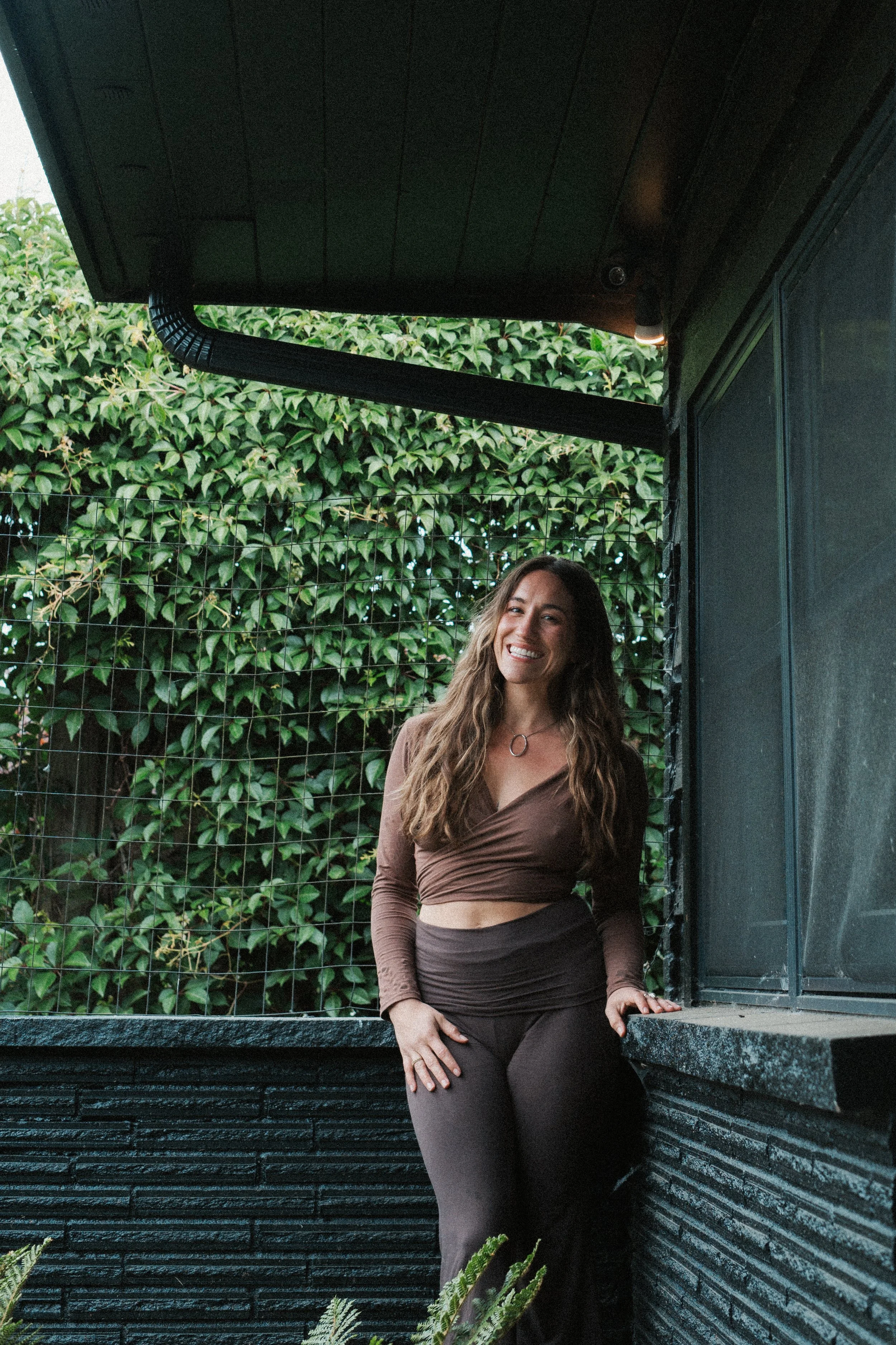 A woman with long wavy hair wearing a brown long-sleeve crop top and dark pants standing outdoors in front of lush greenery and a dark-colored wall, smiling at the camera.