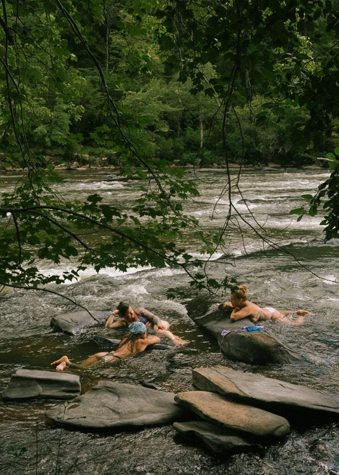 Three people swimming and lounging on large rocks in a river surrounded by green trees.