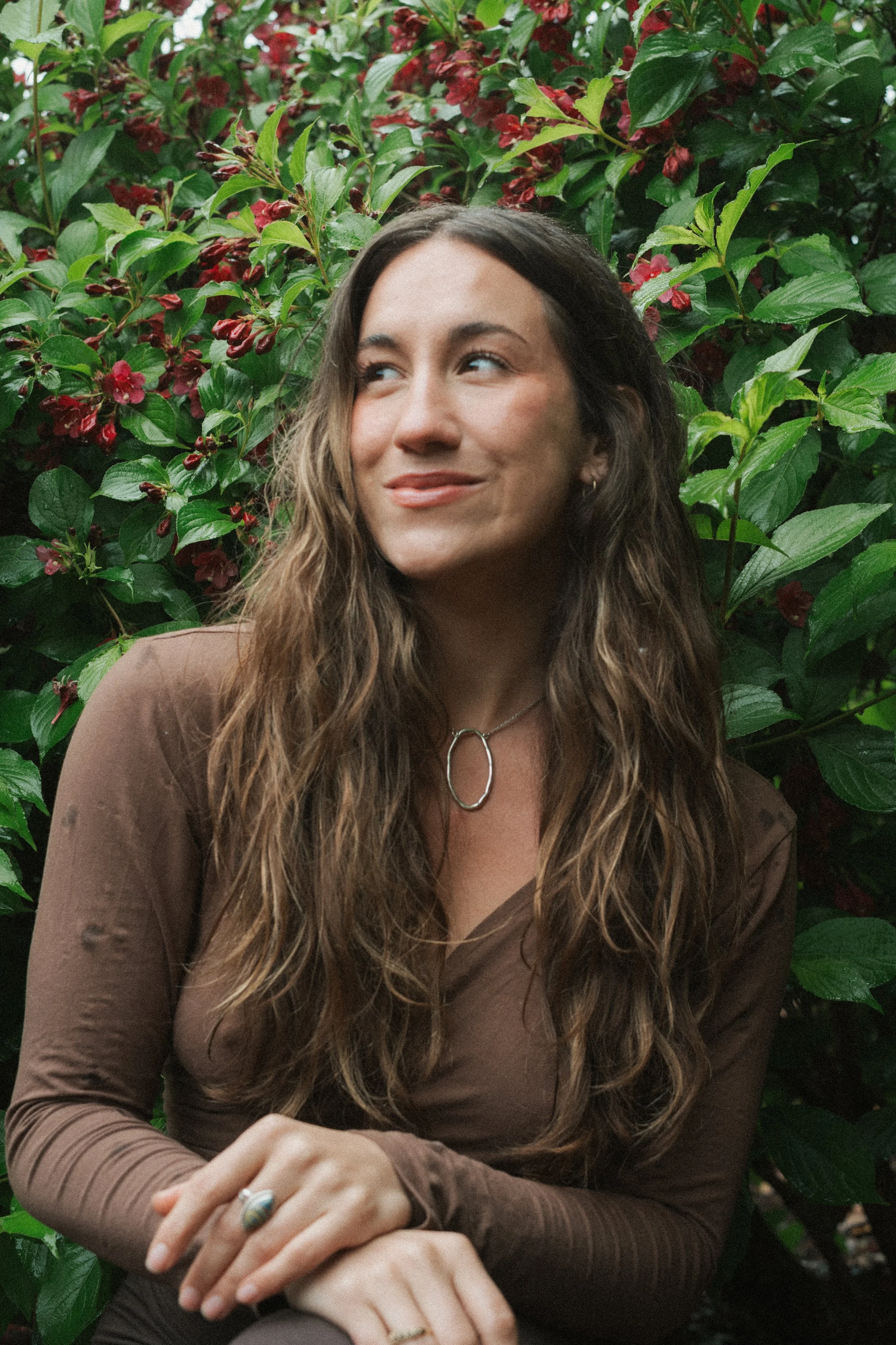 A woman with long wavy brown hair and light skin, wearing a brown top and a silver necklace with a circular pendant, is sitting outdoors in front of a lush green and red flowering bush. She is smiling softly and looking to her right, with her hands resting on her lap.