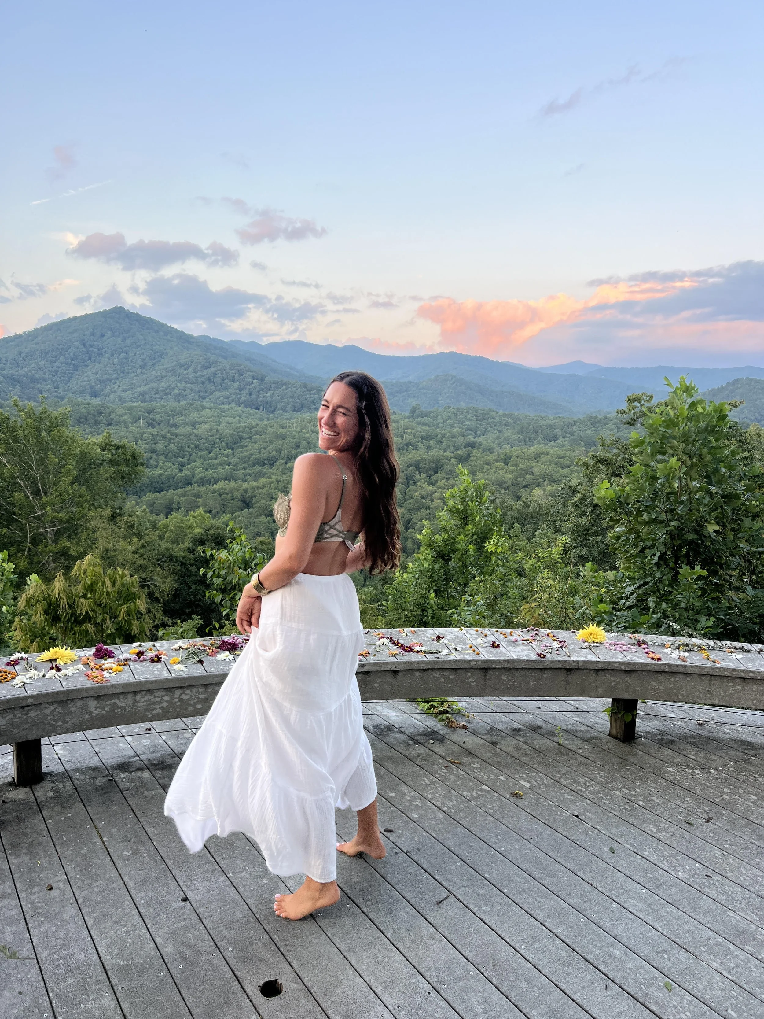 A woman in a white dress is smiling and looking over her shoulder while standing barefoot on a wooden deck with a nature and mountain landscape in the background during sunset.
