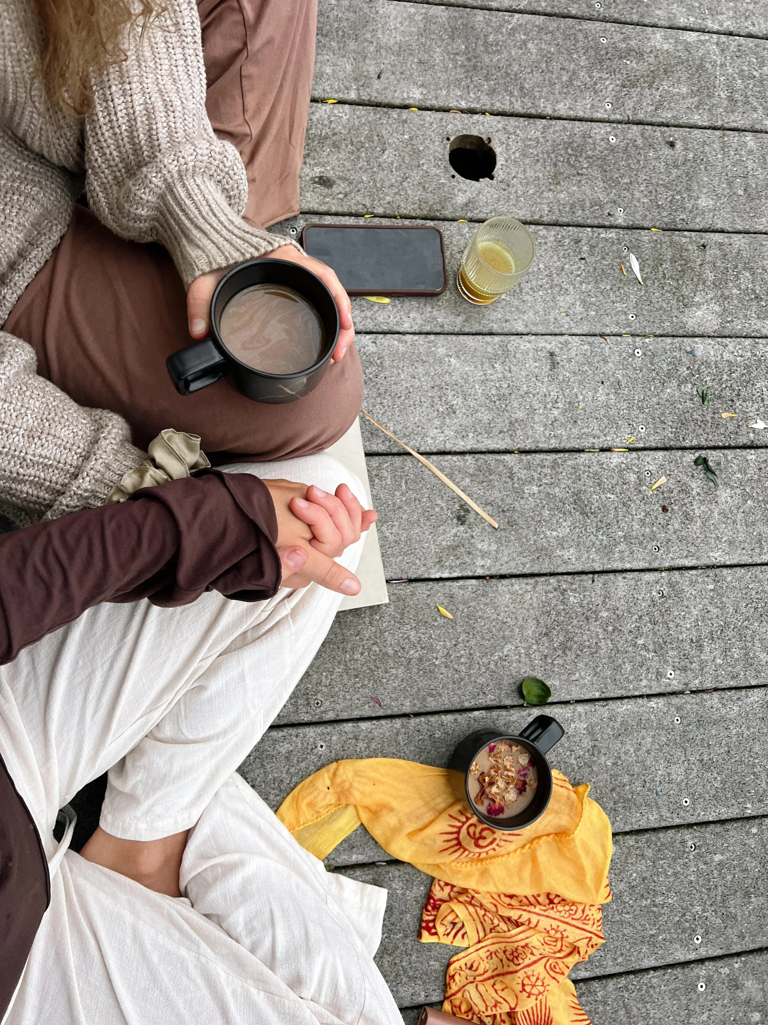 View of people sitting on a wooden deck, with one person holding a mug of tea or coffee, and two other cups of drinks and a bowl of food on the ground. The scene includes a smartphone, a yellow cloth, and a few scattered leaves and petals.