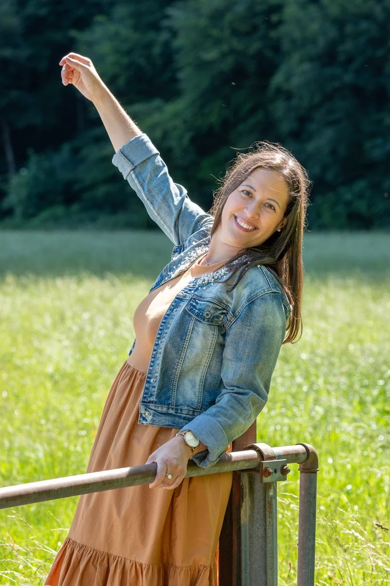 Lächelnde Mentaltrainerin Elke Kieber in einem grünen Feld, trägt einen Jeansjacke und eine orangefarbene Rock, steht am Zaun und hebt den Arm in die Luft, bei Sonnenschein.