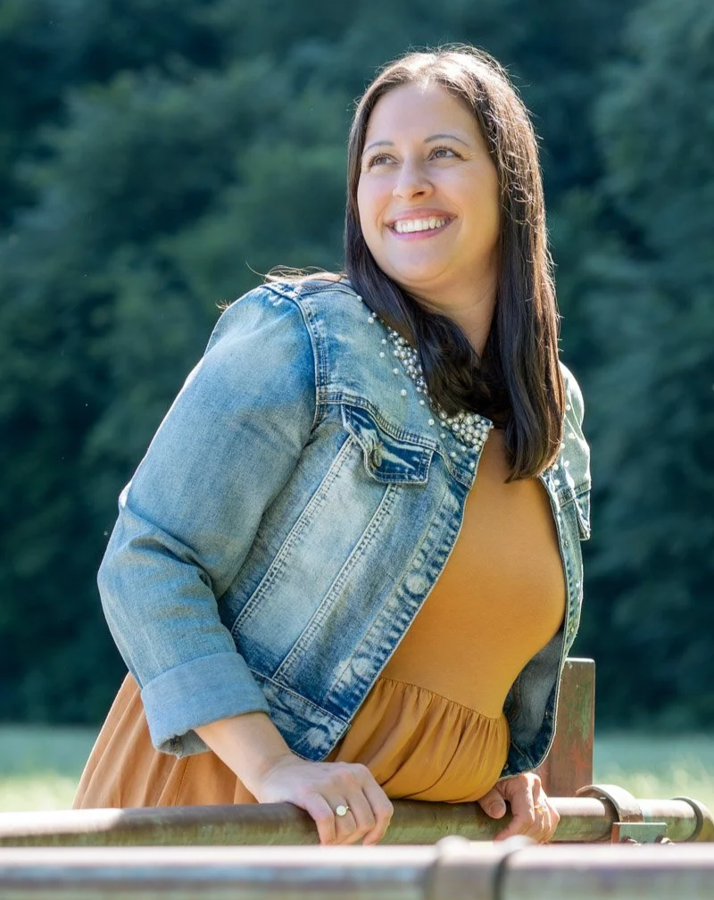 Lächelnder Life Coach Elke Kieber im Freien trägt eine Jeansjacke und ein gelbes Kleid, lehnt an einem Klettergerüst, im Hintergrund Bäume.