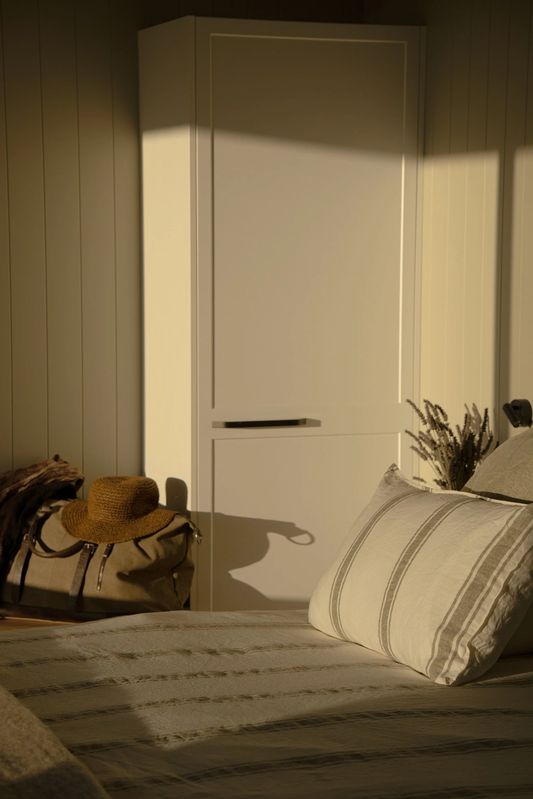 A bedroom scene with a bed covered in a striped bedspread and pillows, a white wardrobe, a dried plant in the corner, and a hat placed on a bag near the foot of the bed, with warm lighting.
