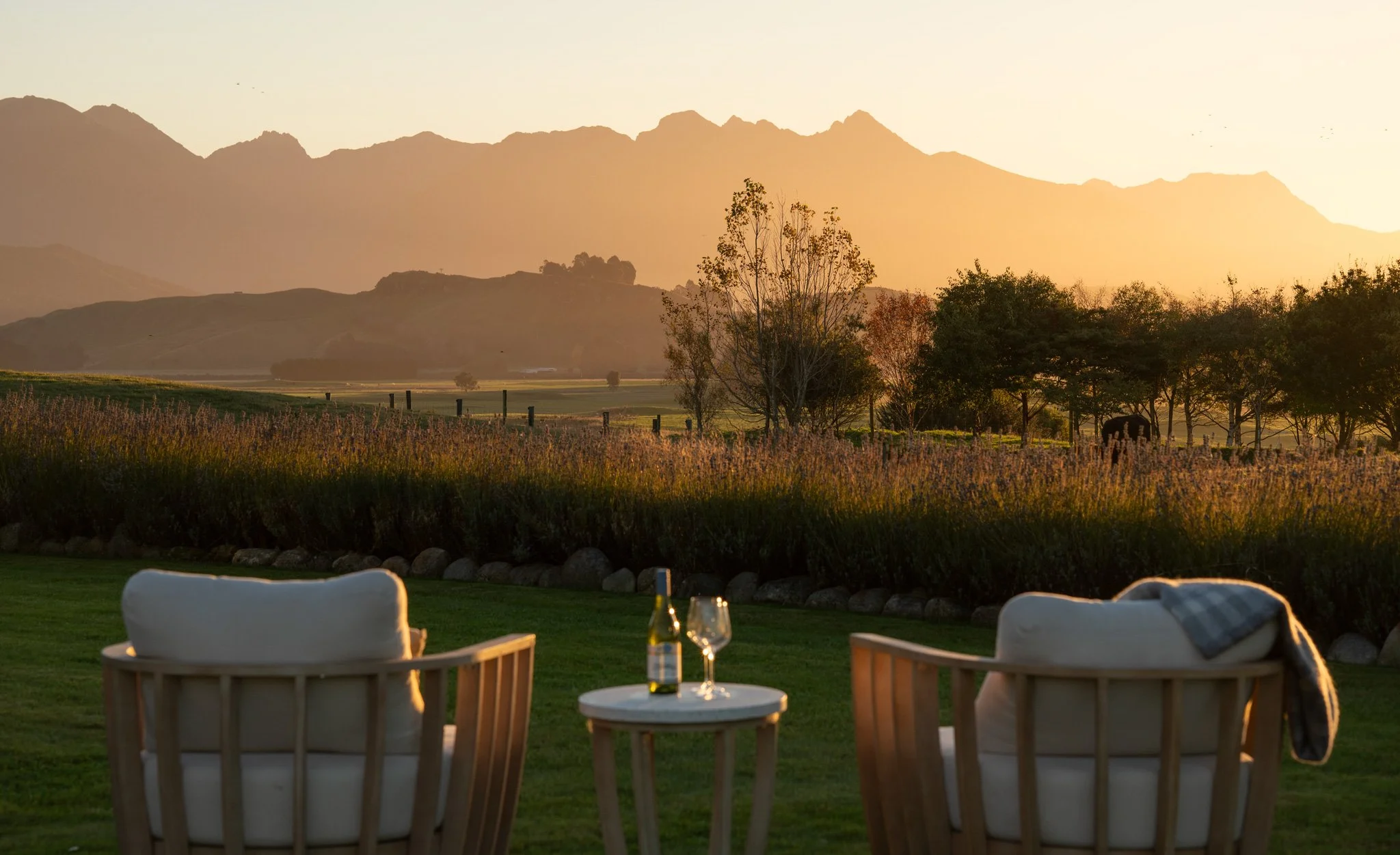 An outdoor setting at sunset with empty chairs and tables in foreground, with trees, a fence, and mountain range in background.