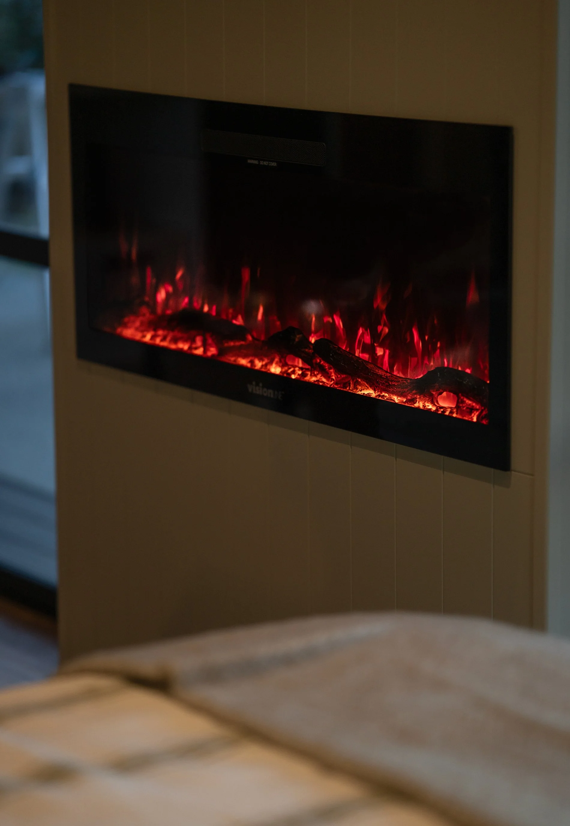 Electric fireplace with glowing flames mounted on white shiplap wall in Maunga Tipu studio, providing warmth and atmosphere for year-round comfort