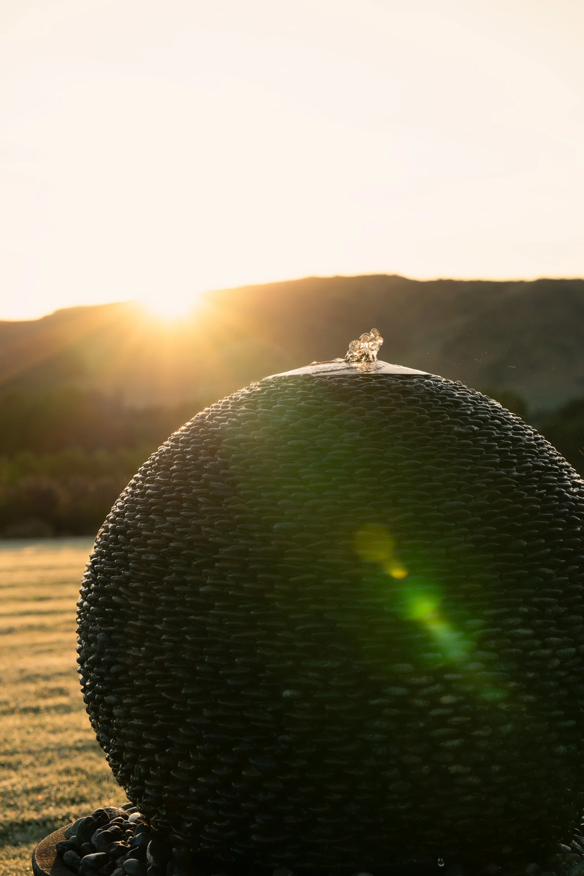 Close-up of a black, pebble-covered sculpture or fountain with water splashing at the top, set against a sunset background with hills and a body of water.