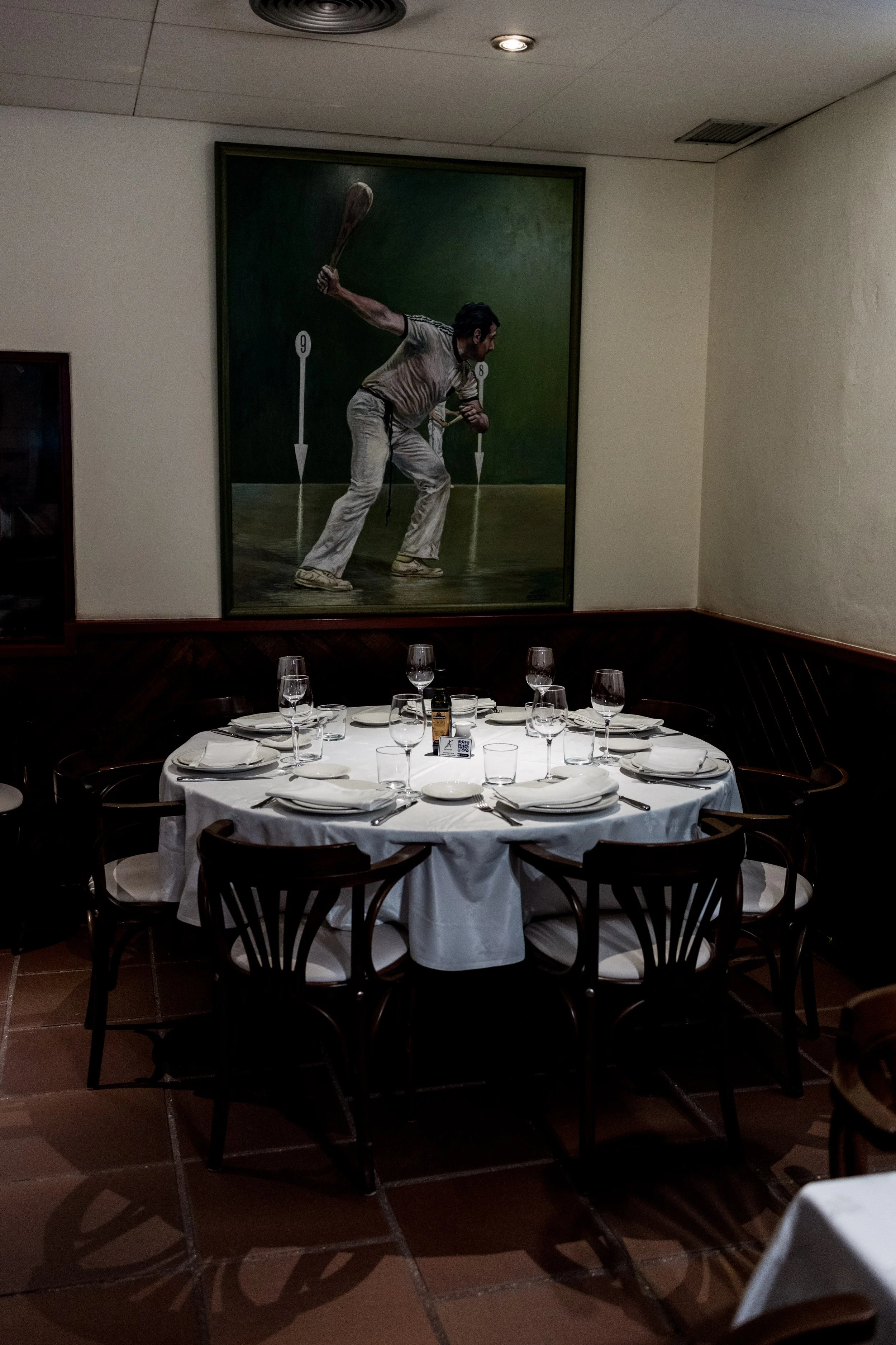 Detalle de una mesa decorada con mantel blanco, platos, cubiertos, vasos y copas en un restaurante. En la pared, un mural que muestra un jugador de tenis en acción.
