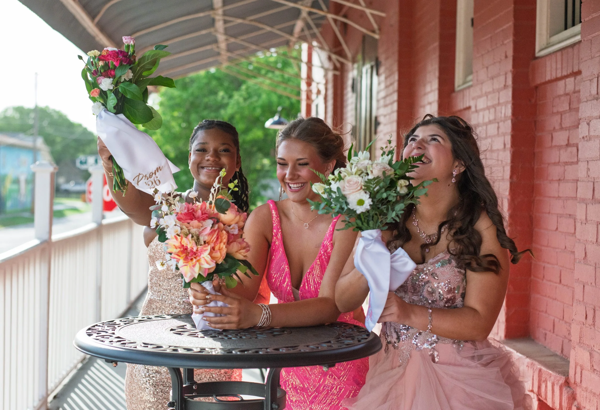 Three women celebrating on a porch, holding bouquets of flowers, dressed in formal and colorful outfits, smiling and laughing.