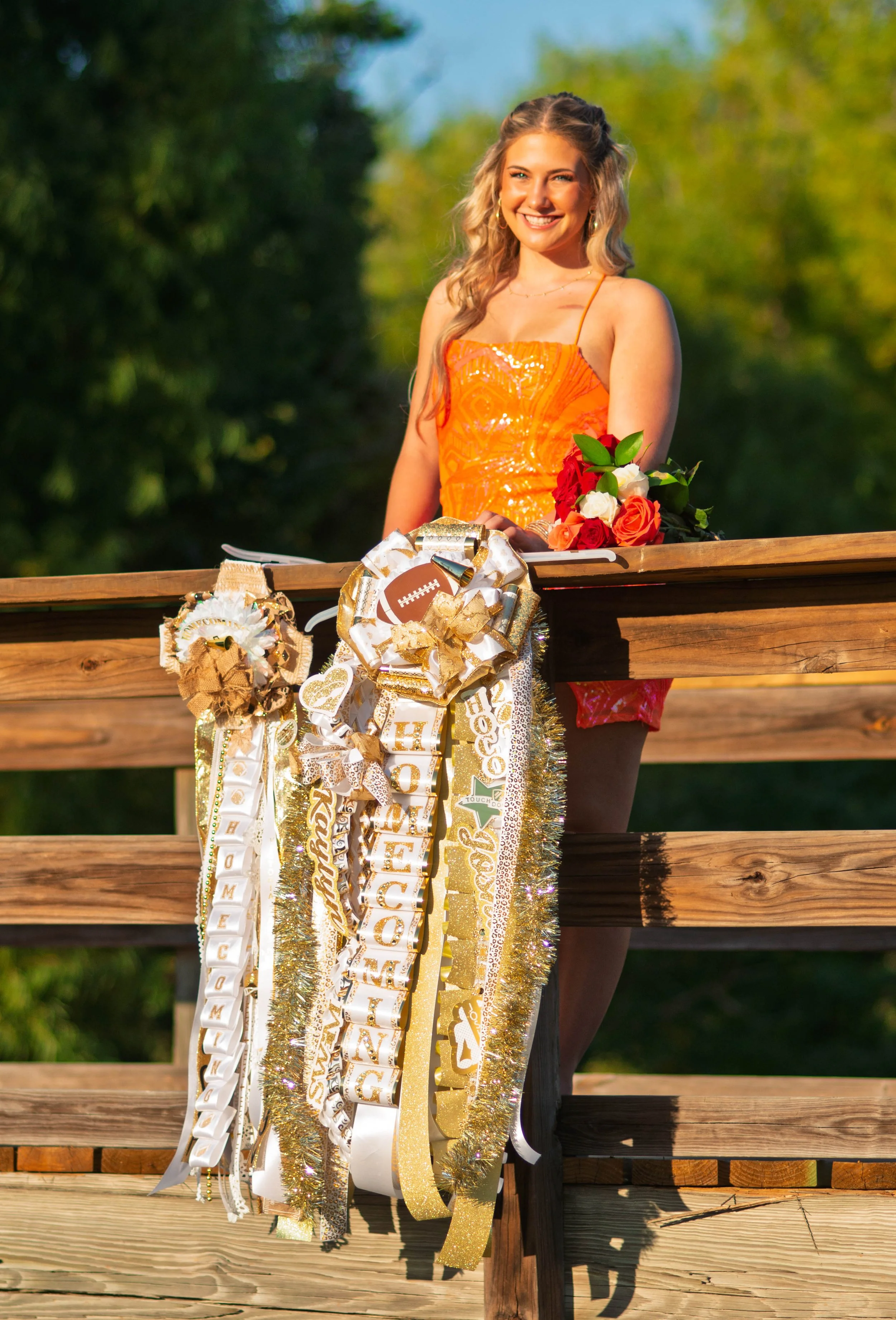 Young woman smiling, wearing an orange top, with floral accessories, standing behind a large display of decorated ribbons and flowers on a wooden ledge outdoors.