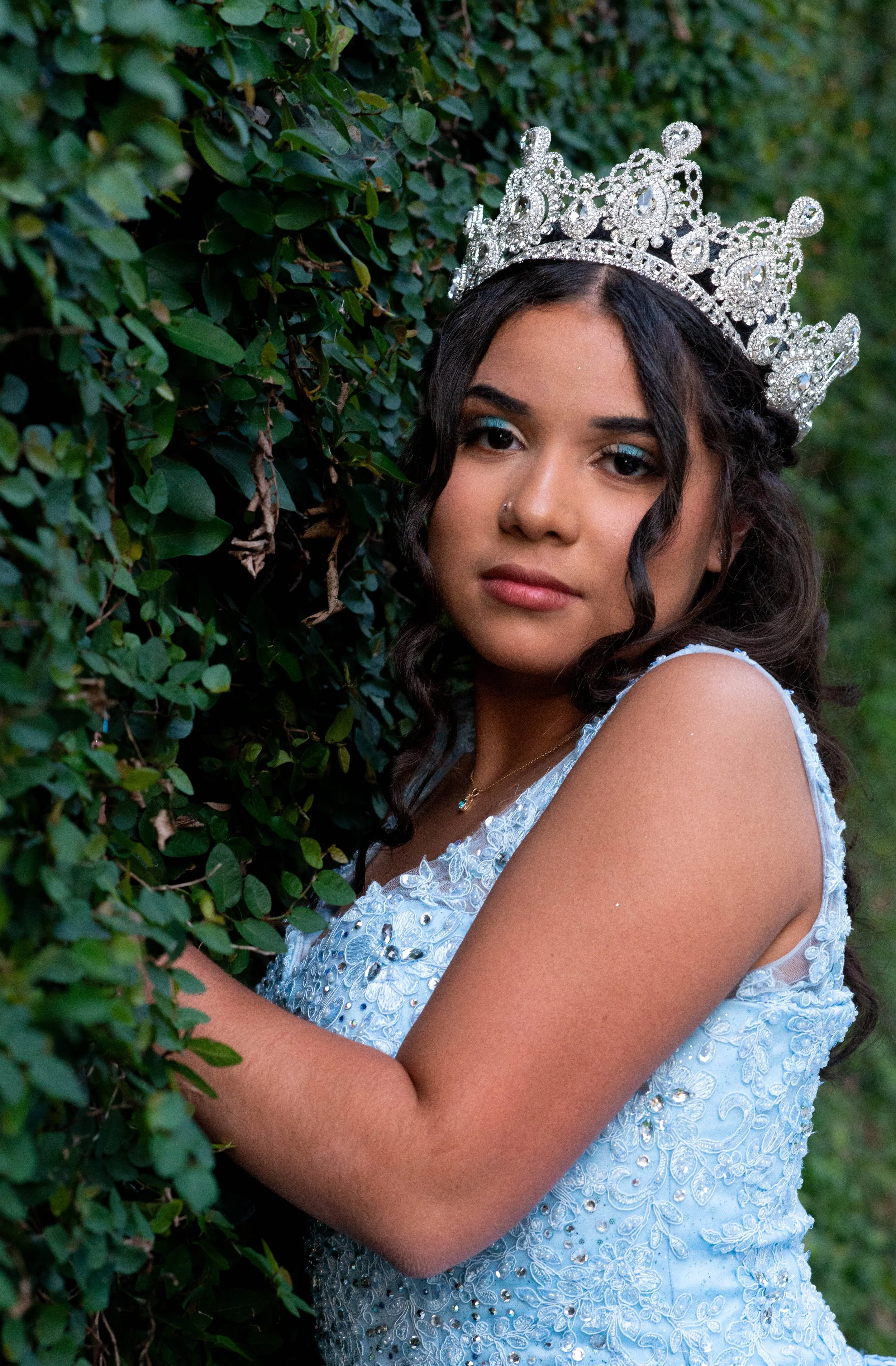 Young woman wearing a silver tiara and a light blue embroidered dress, standing beside a bush, during daytime.