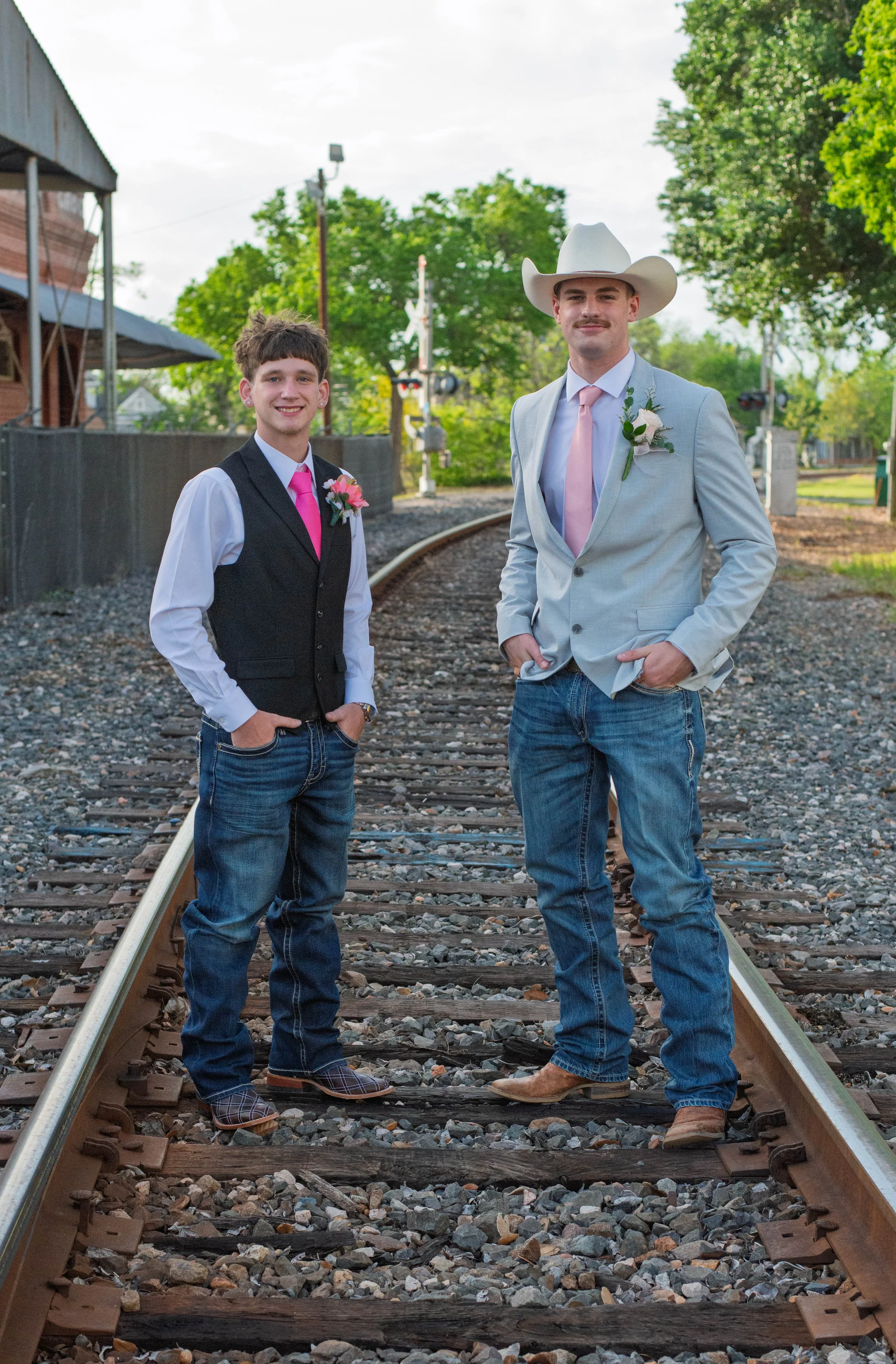 Two young men stand on train tracks, dressed in formal attire with pink accents, smiling at the camera outdoors.