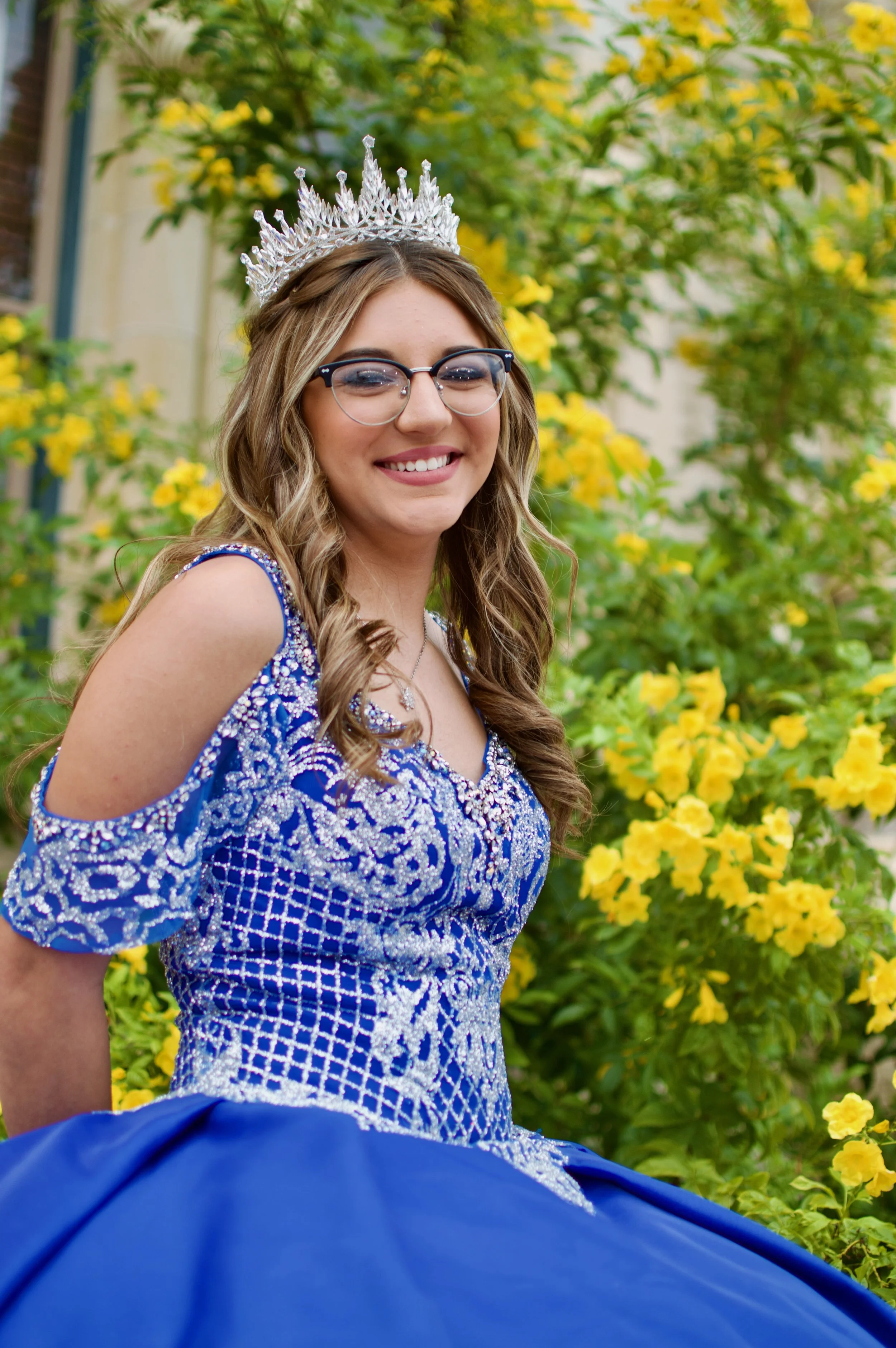 Young woman wearing a blue and silver embellished gown and a tiara, smiling outdoors with yellow flowers in the background.