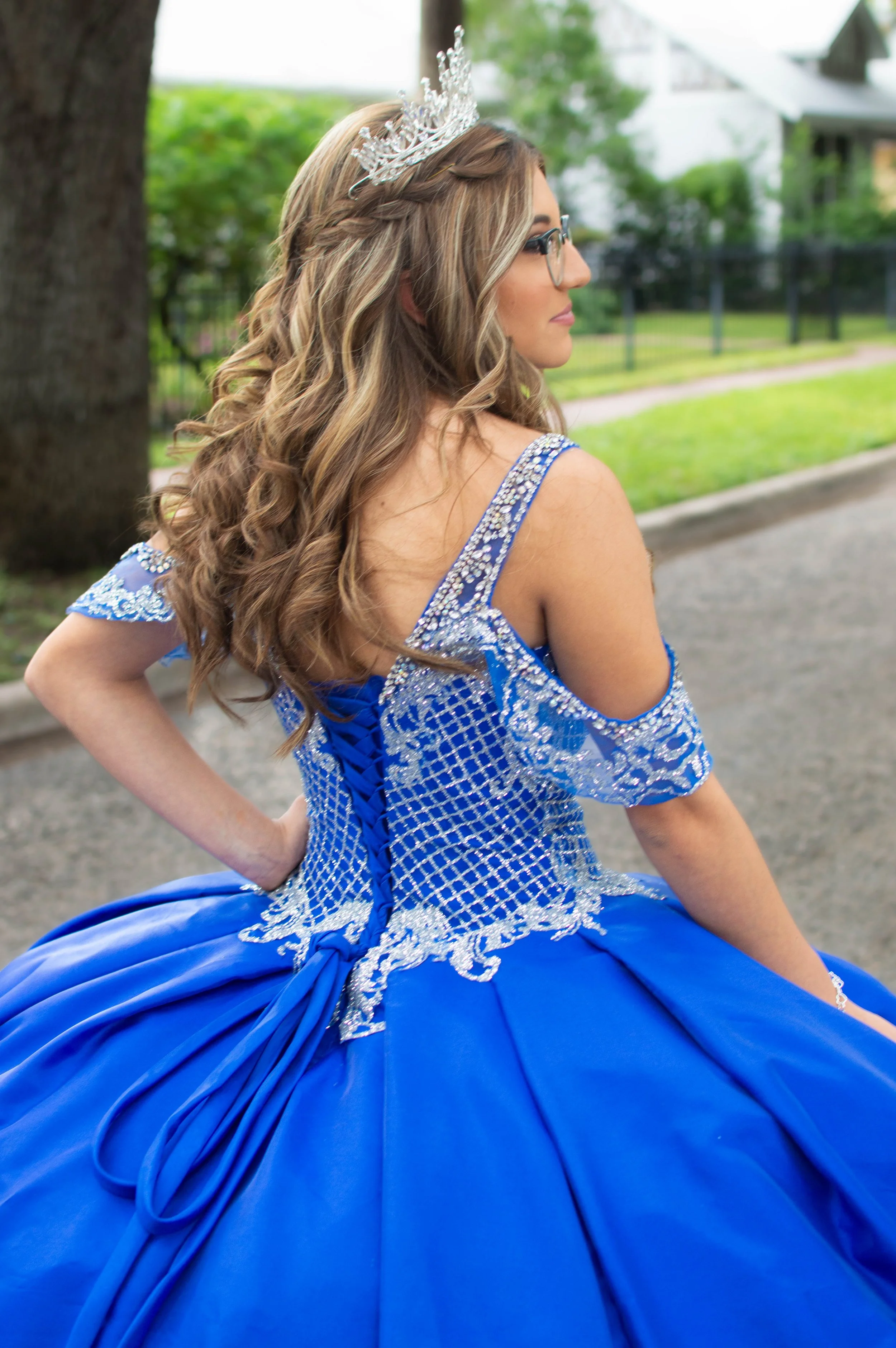 A young woman in a royal blue quinceañera dress with silver embellishments, wearing glasses, a bracelet, and a tiara, sitting outdoors in a park with trees and grass.