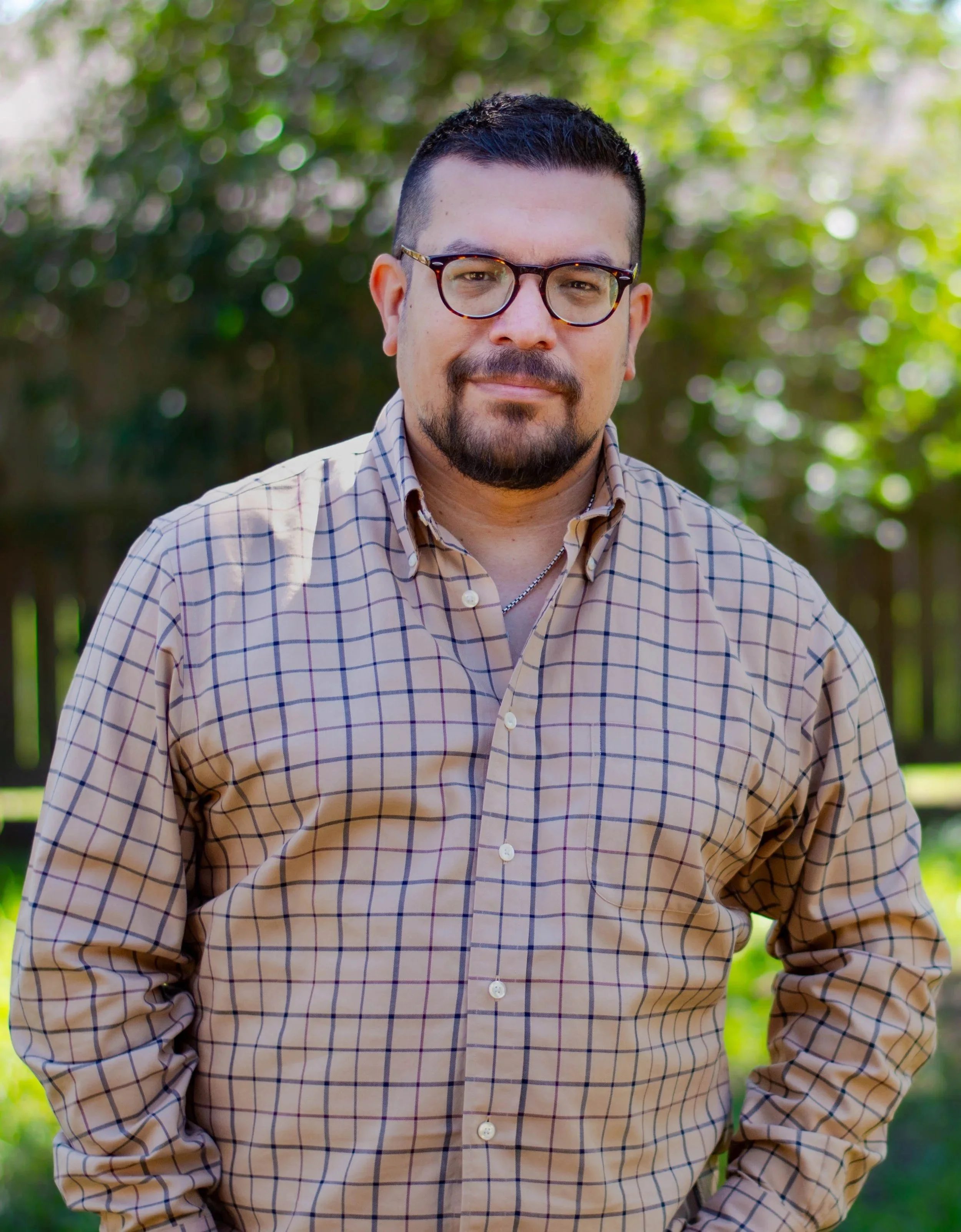 Man wearing glasses and a beige checkered shirt standing outdoors with green foliage in the background.