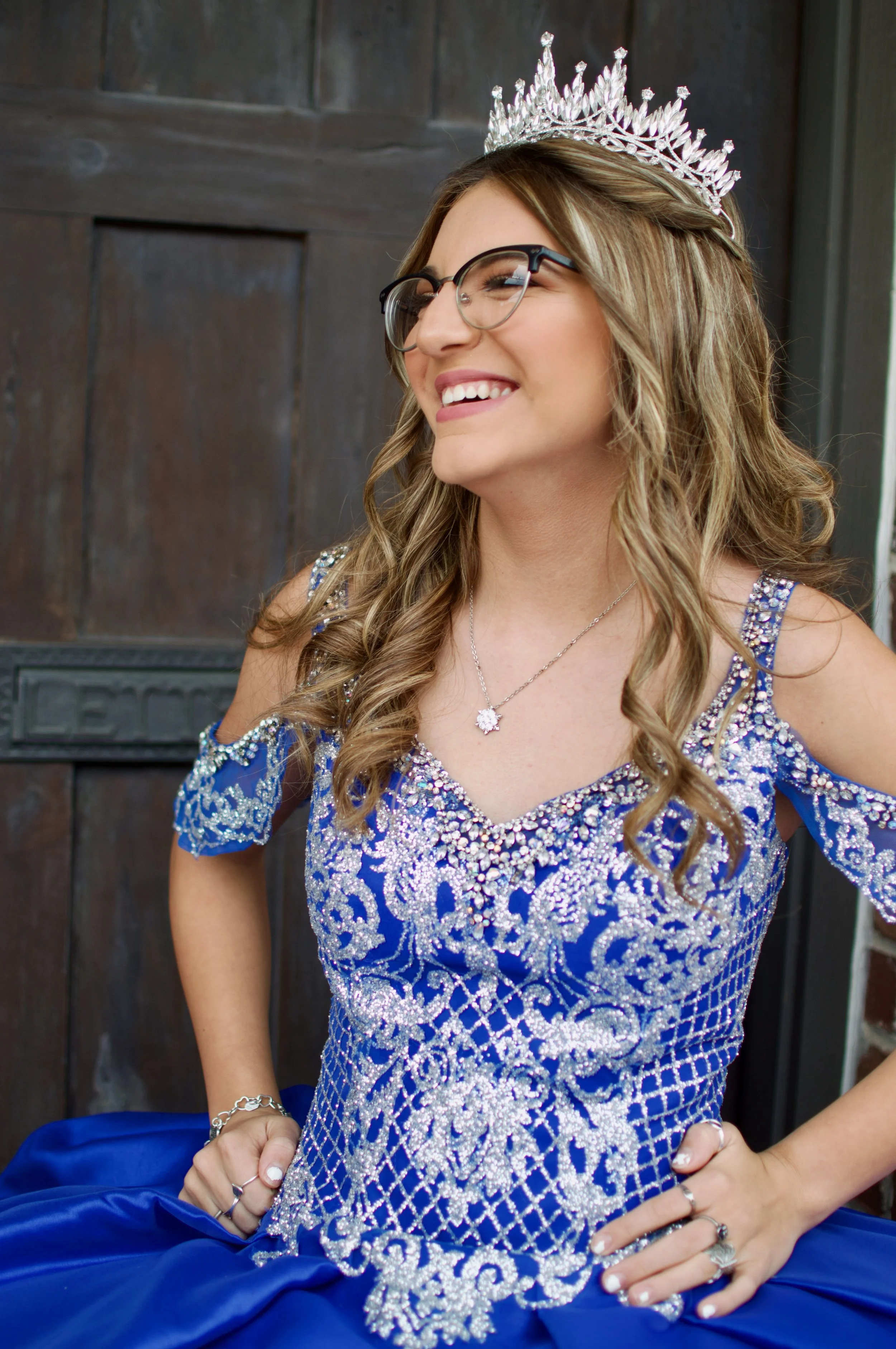A young woman wearing glasses, a tiara, and a royal blue dress with elaborate silver and rhinestone embroidery, smiling and looking off to the side.