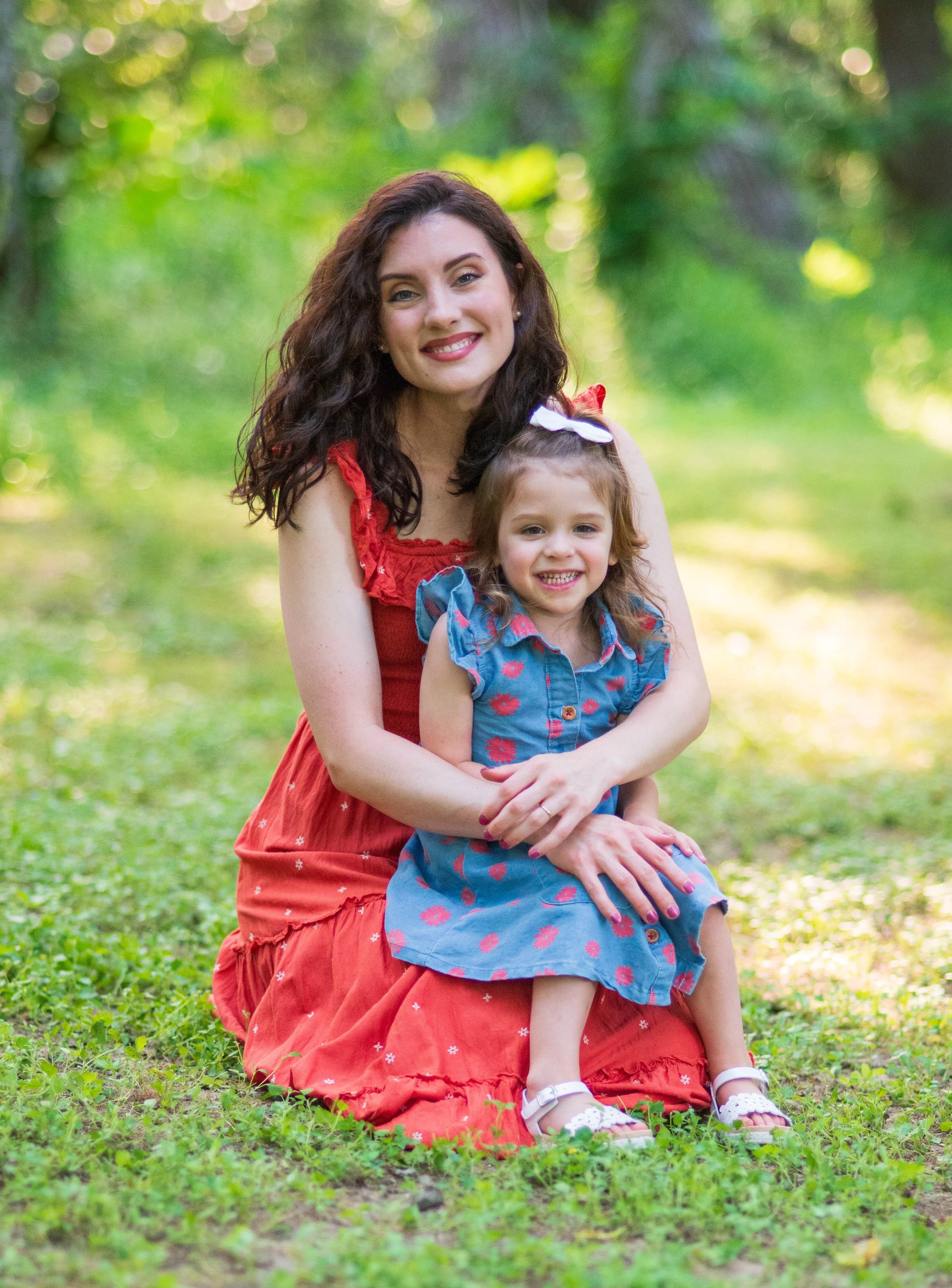 A woman and a young girl sitting on the grass in a sunny, wooded area, smiling at the camera.