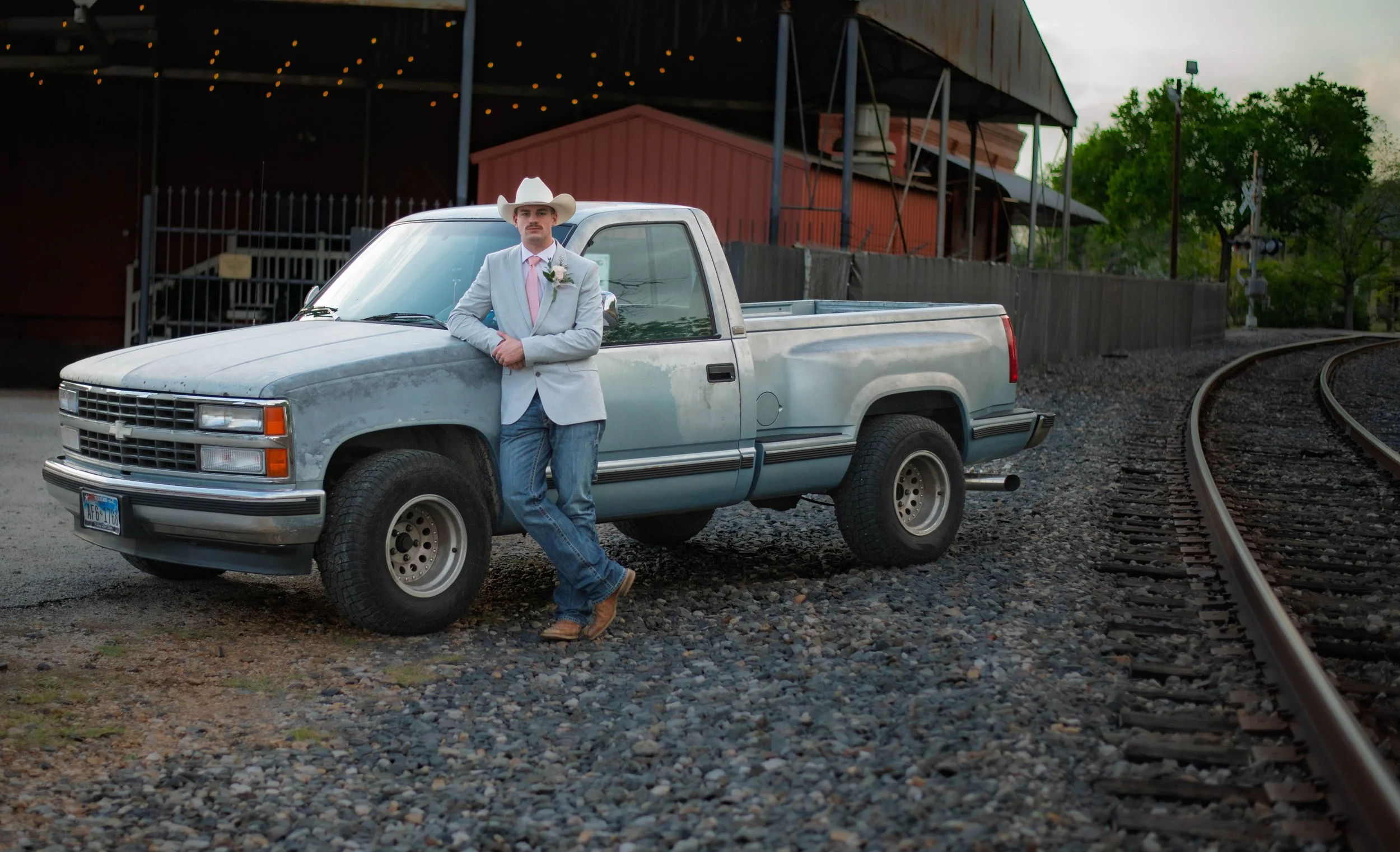 A man in a light gray suit, pink tie, and white cowboy hat leaning against a silver pickup truck on a gravel road beside railroad tracks, with a barn and trees in the background.