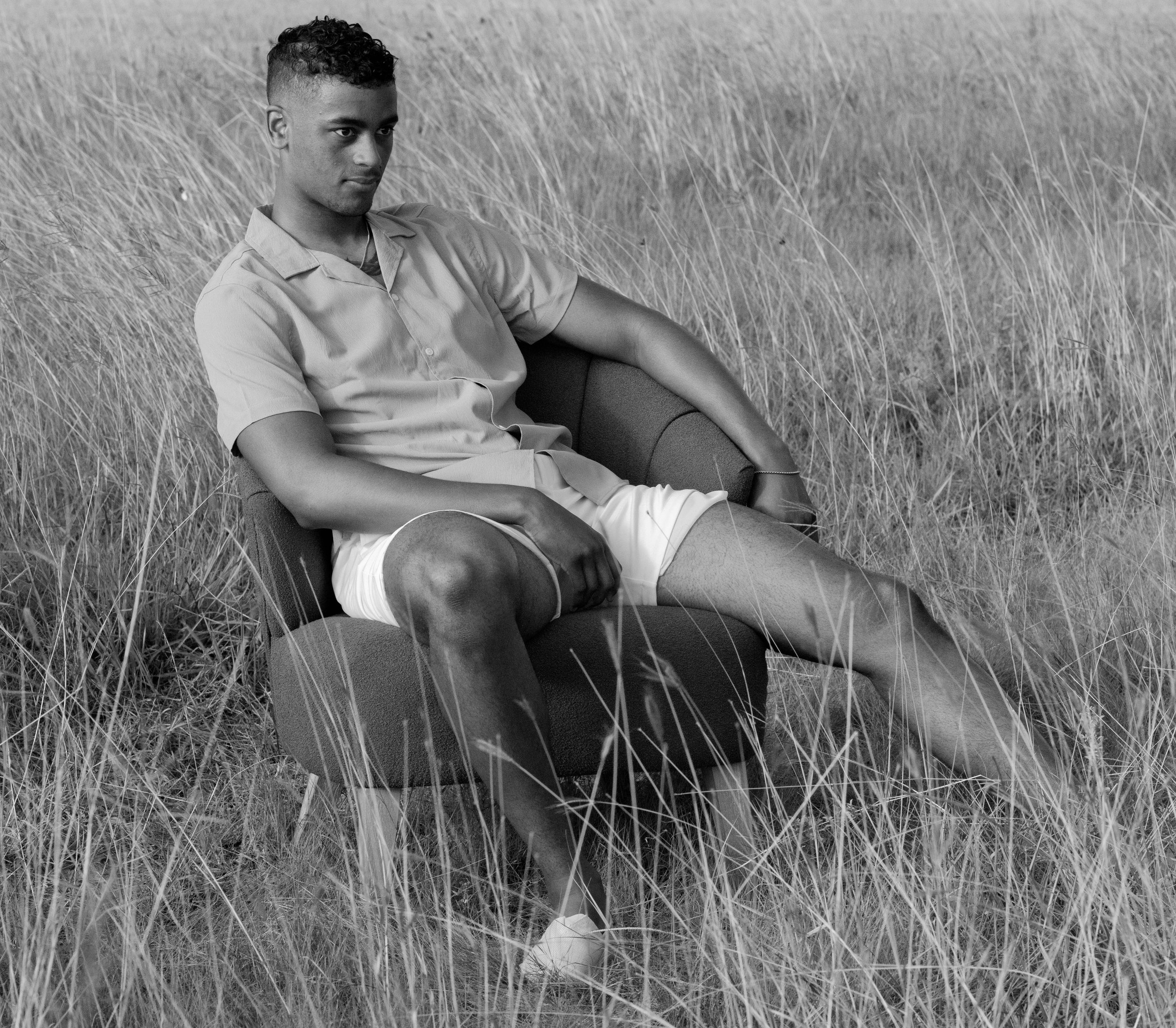 A young man sitting on a chair in a grassy field, wearing a casual button-up shirt and shorts, looking pensively to the side.