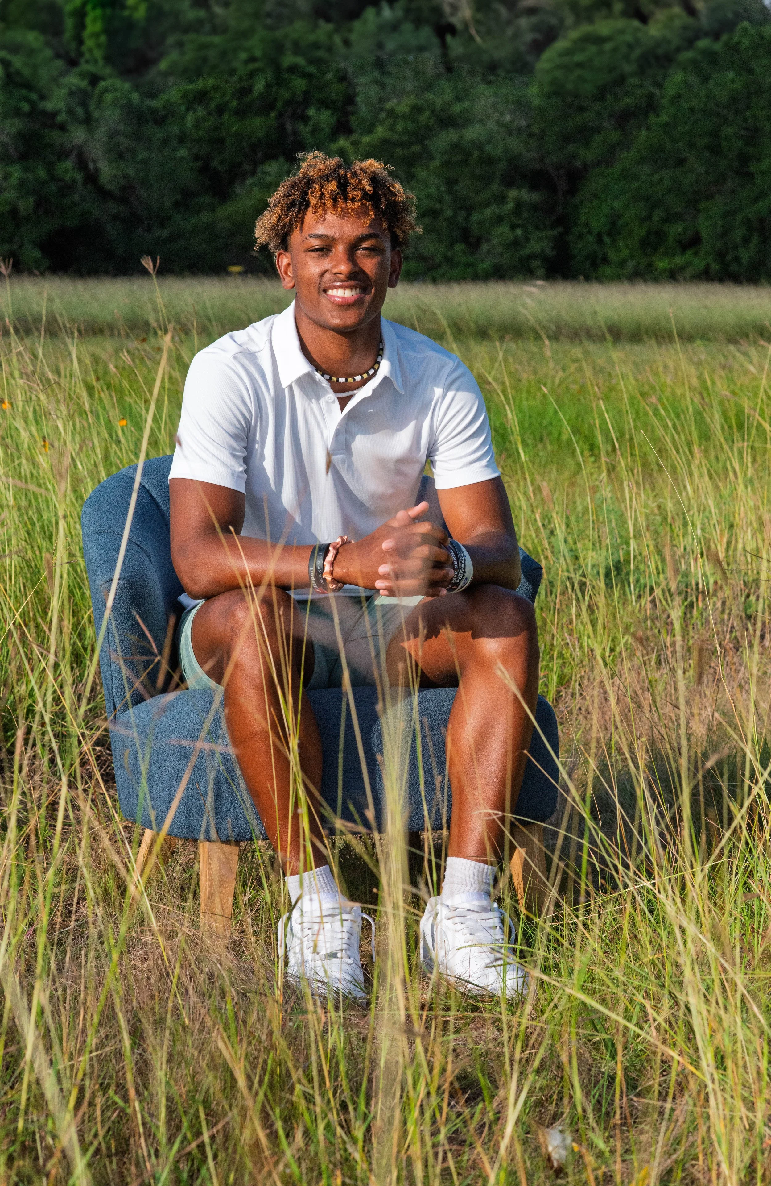 A young man sitting on a blue chair in a field of tall grass with green trees in the background, smiling at the camera.