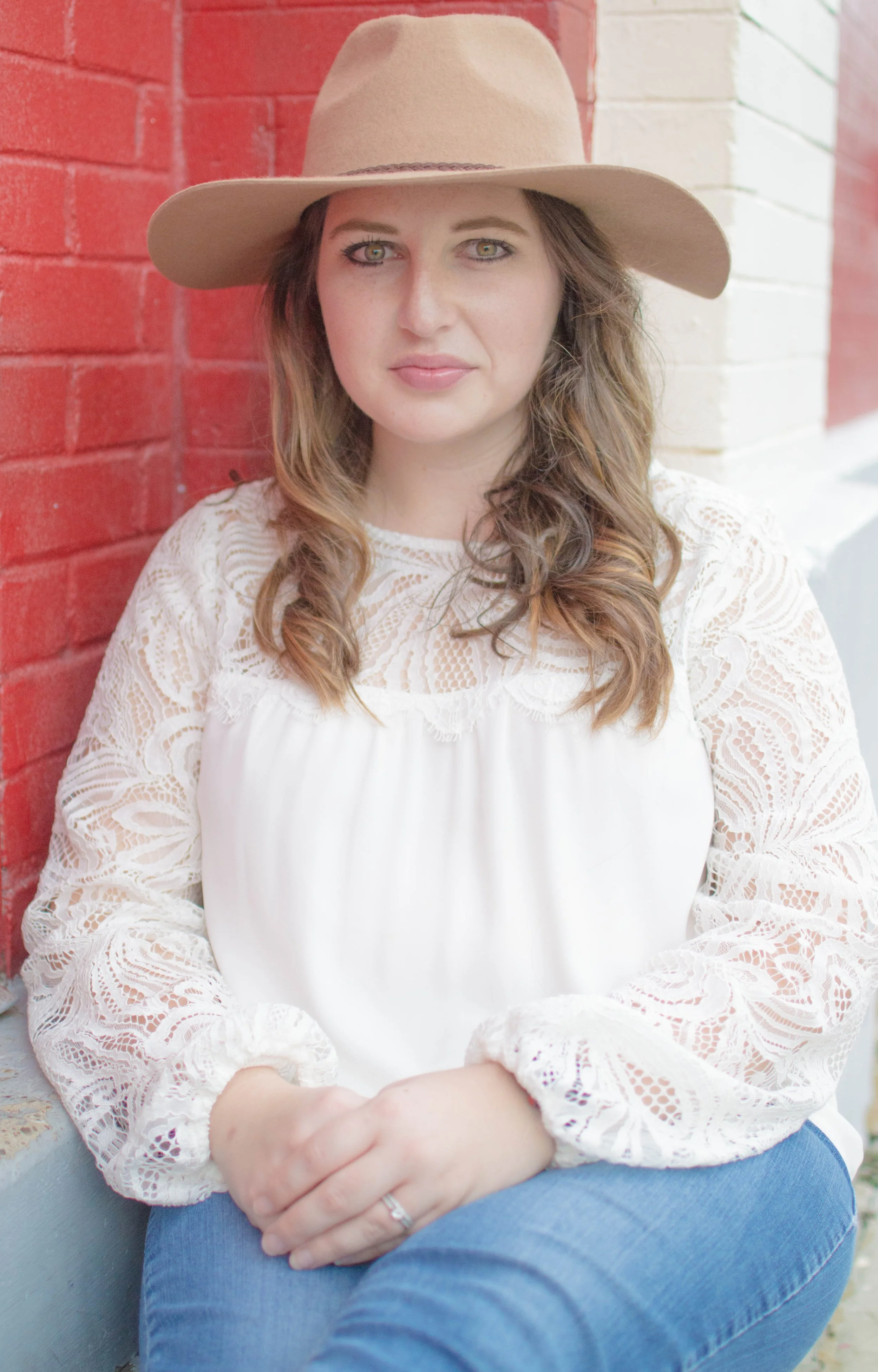 Woman with wavy brown hair wearing a wide-brimmed beige hat, white lace top, and jeans, sitting against a red brick wall