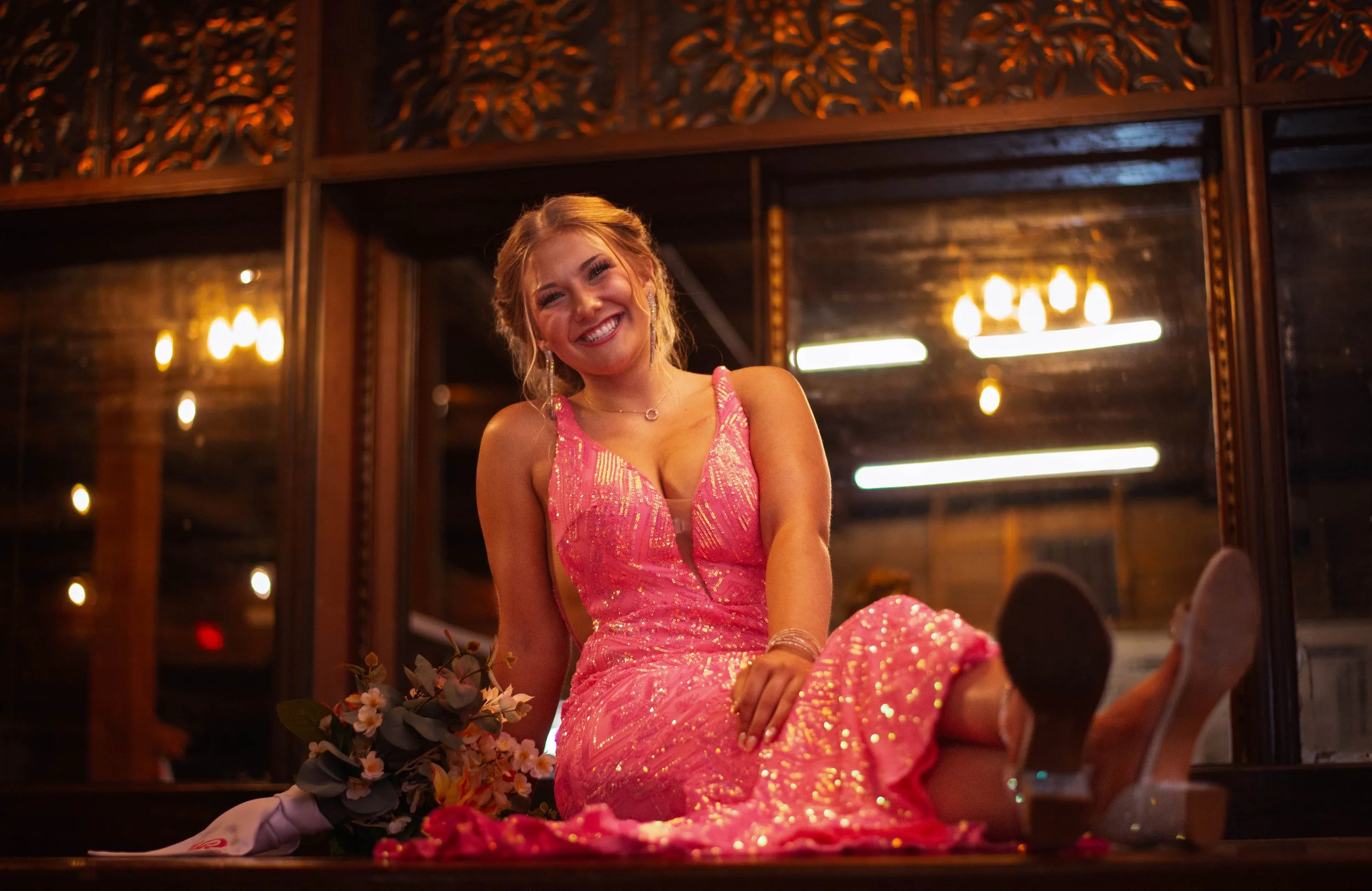 A smiling young woman wearing a pink, glittery dress, sitting on the floor with a bouquet of flowers beside her in a warmly lit room with decorative wood paneling and warm yellow lights reflected in large mirrors.