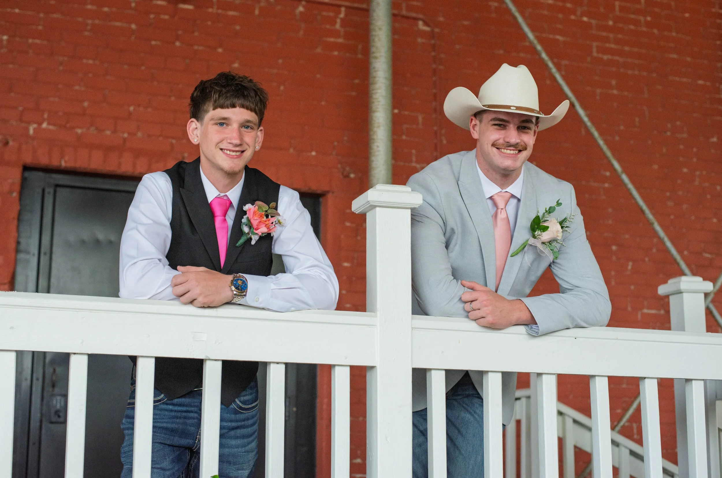 Two young men dressed in suits and ties, with boutonnières, standing behind a white railing in front of a brick wall, smiling at the camera. One wears a cowboy hat.