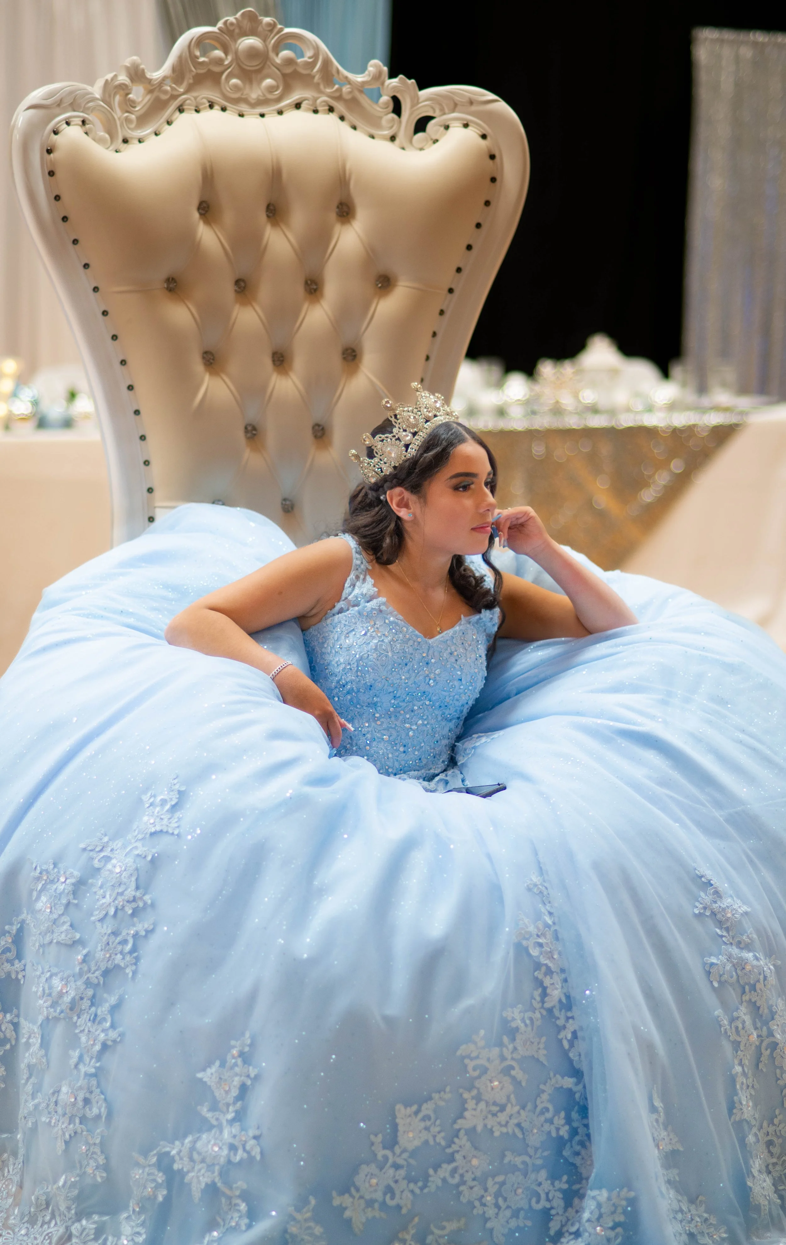 Young woman wearing a blue gown and crown, sitting on a large, ornate bed with a high, tufted headboard, in a decorated indoor setting.