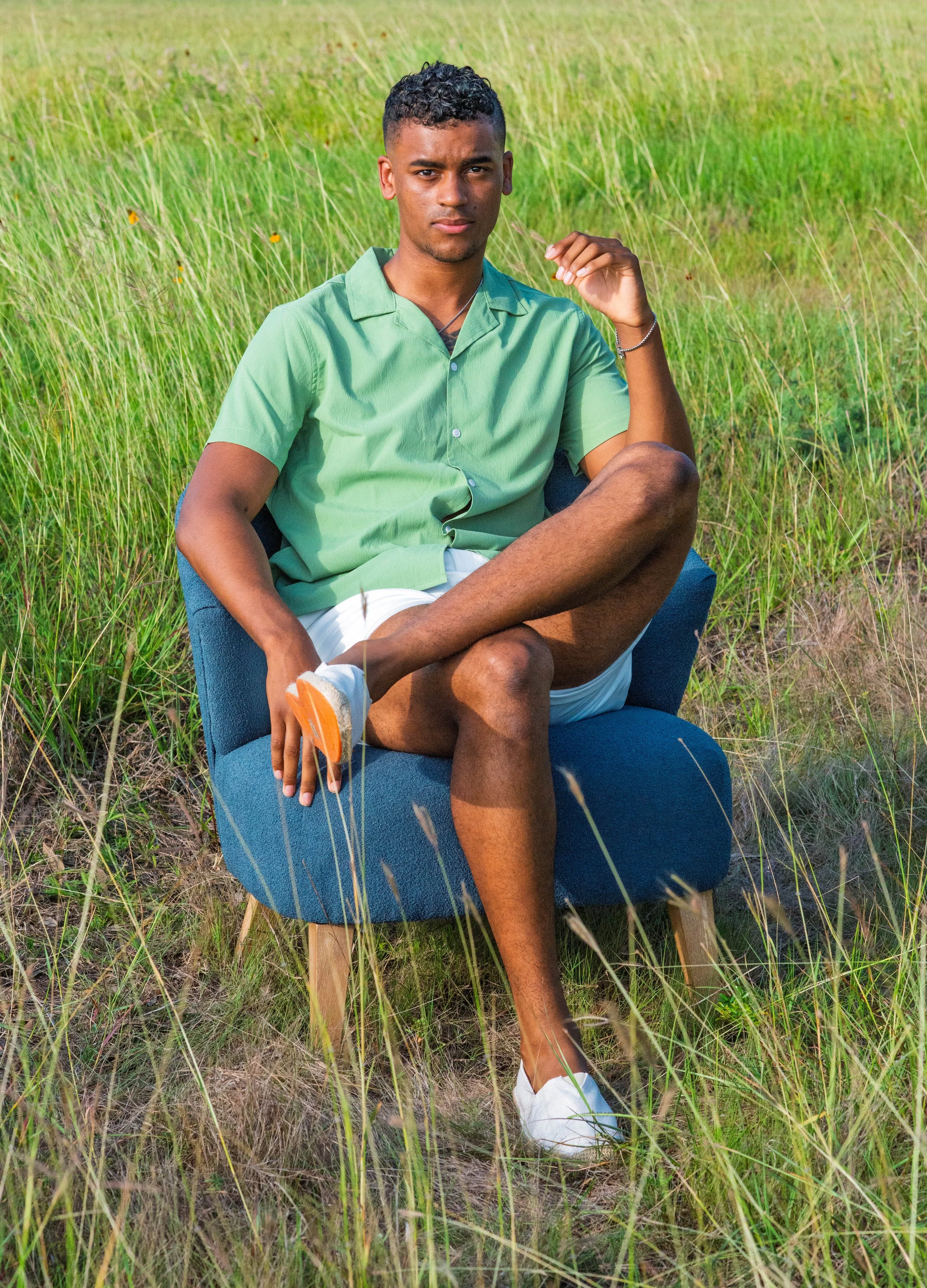 A young man sitting cross-legged on a blue chair in a grassy field, wearing a green shirt, white shorts, and white shoes, holding a frisbee in his right hand, looking at the camera.