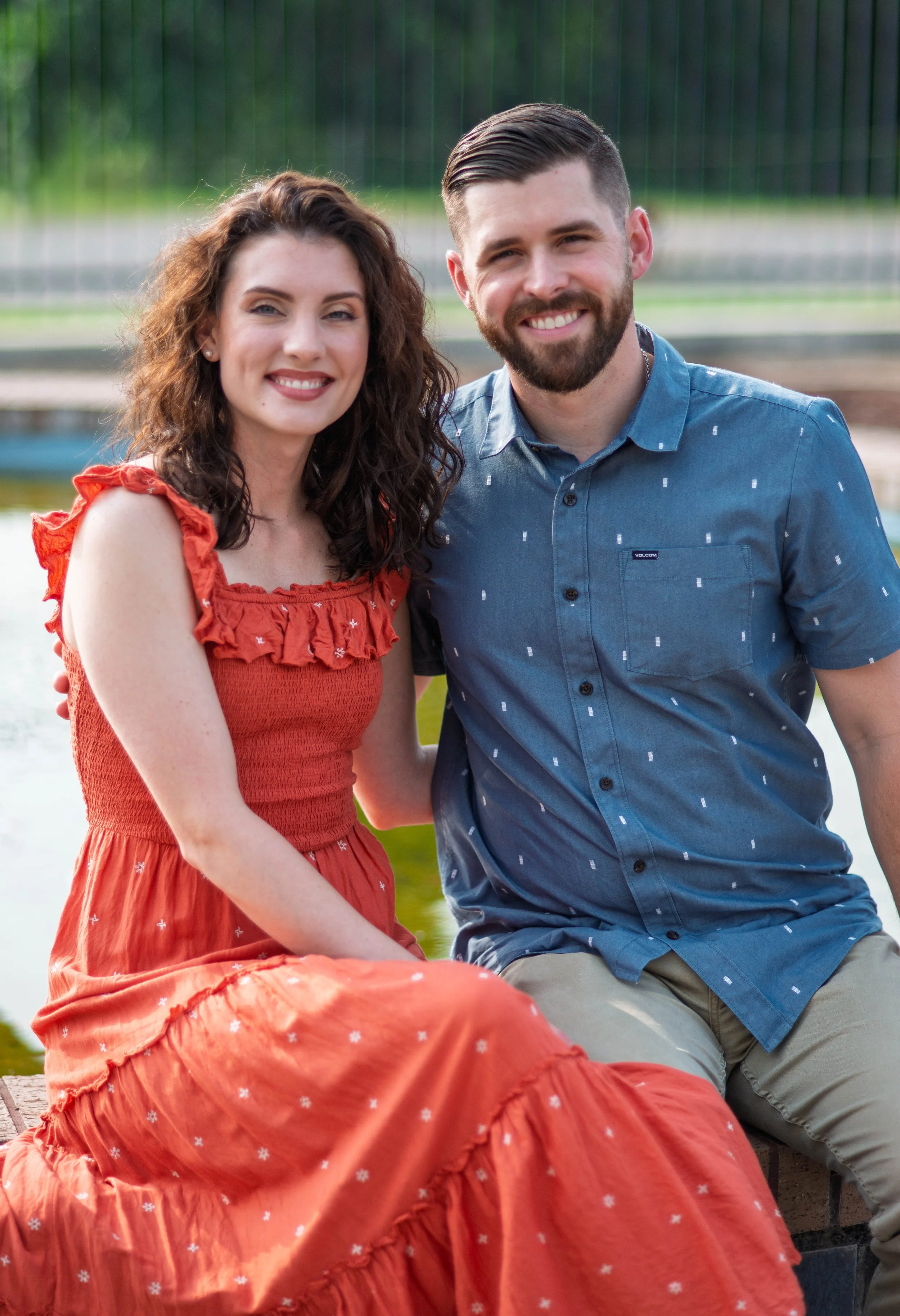 A smiling man and woman sitting closely together outdoors with a body of water and green trees in the background.