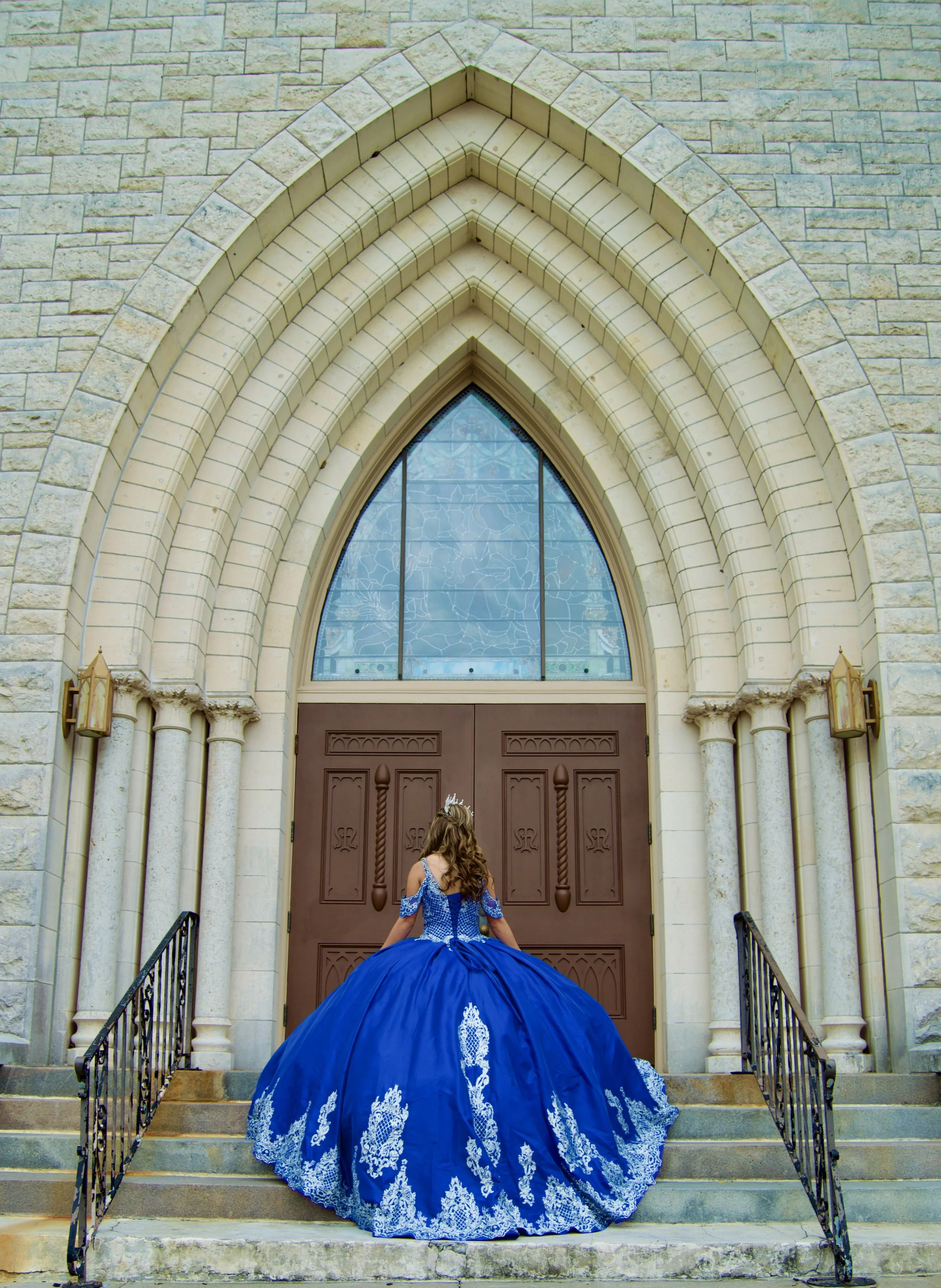 A girl in a royal blue gown with white embroidery, facing a large wooden church door, at the top of stone steps with black iron railings, under an arched stone entrance.