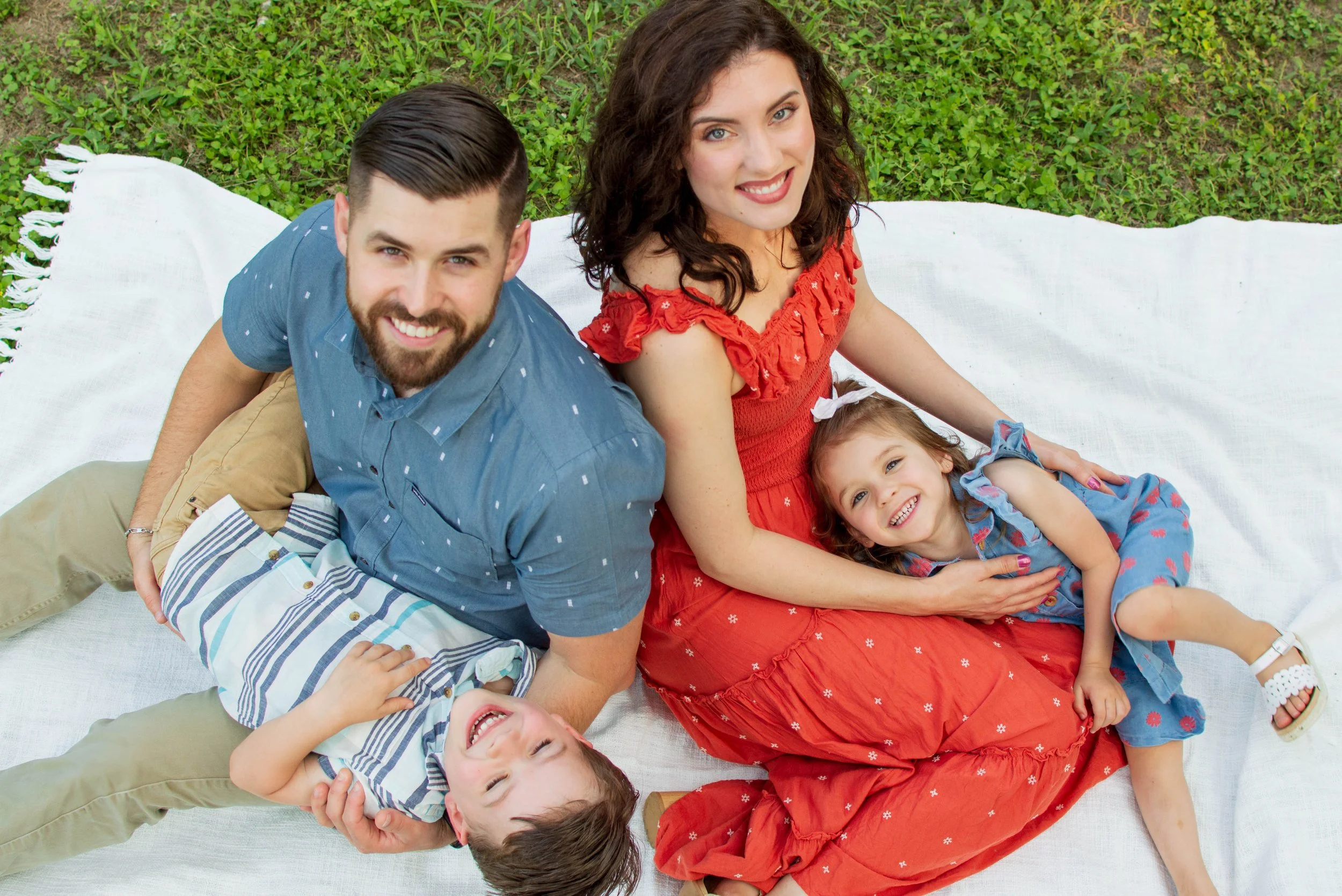 A family of four lying on a white blanket on green grass, smiling and looking up at the camera. The mother wears a red dress, the father a blue shirt, and two young children are with them, one child is in striped shirt and khaki pants, and the other 