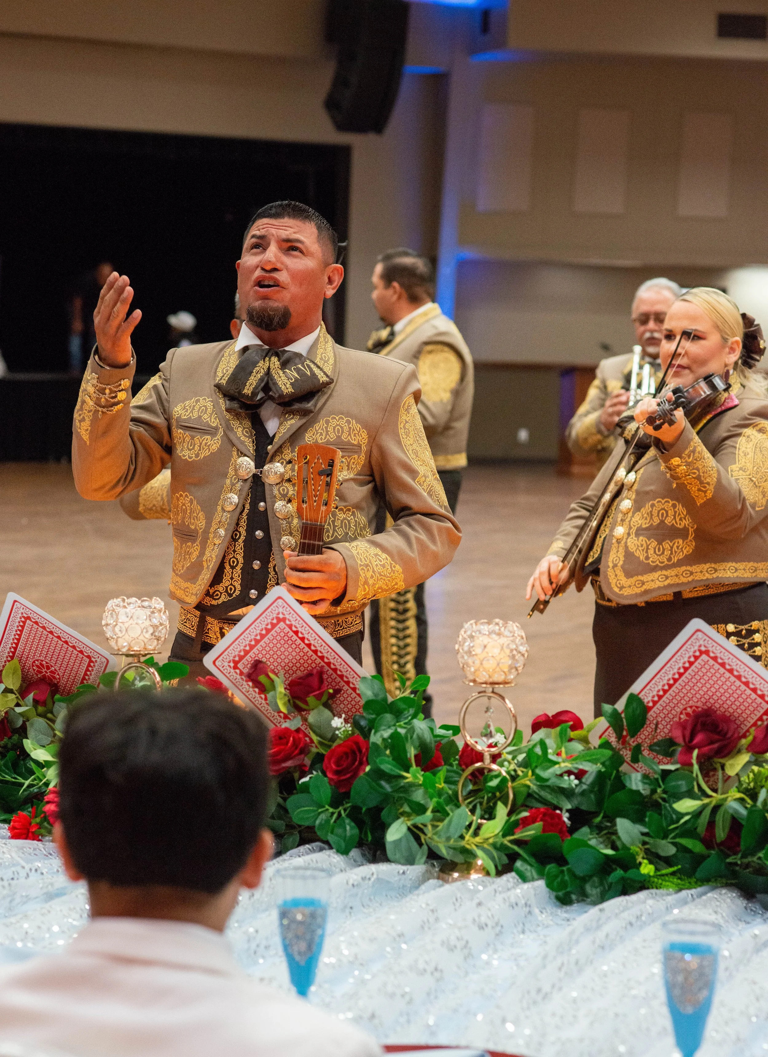 A mariachi band performing at a celebration with ornate costumes, floral decorations, and playing instruments such as guitar and violin.