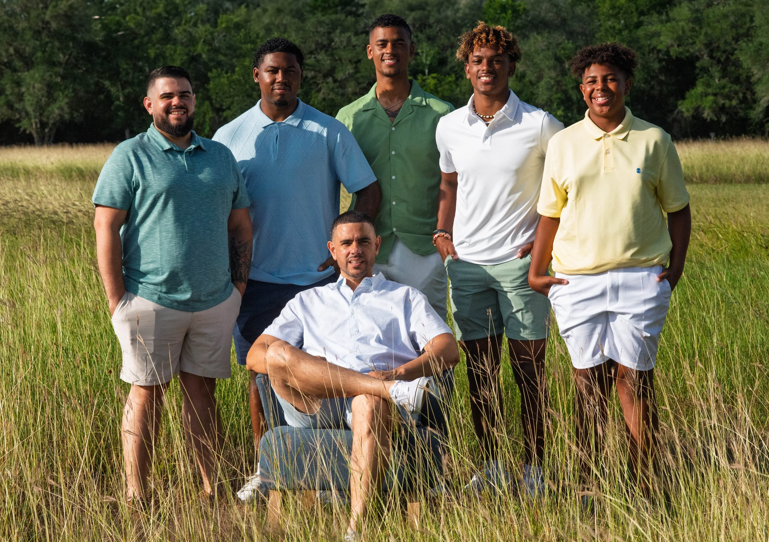 Group of seven young men in casual clothing outdoors in a grassy field with trees in the background. One man is seated in a chair while the others stand around him.