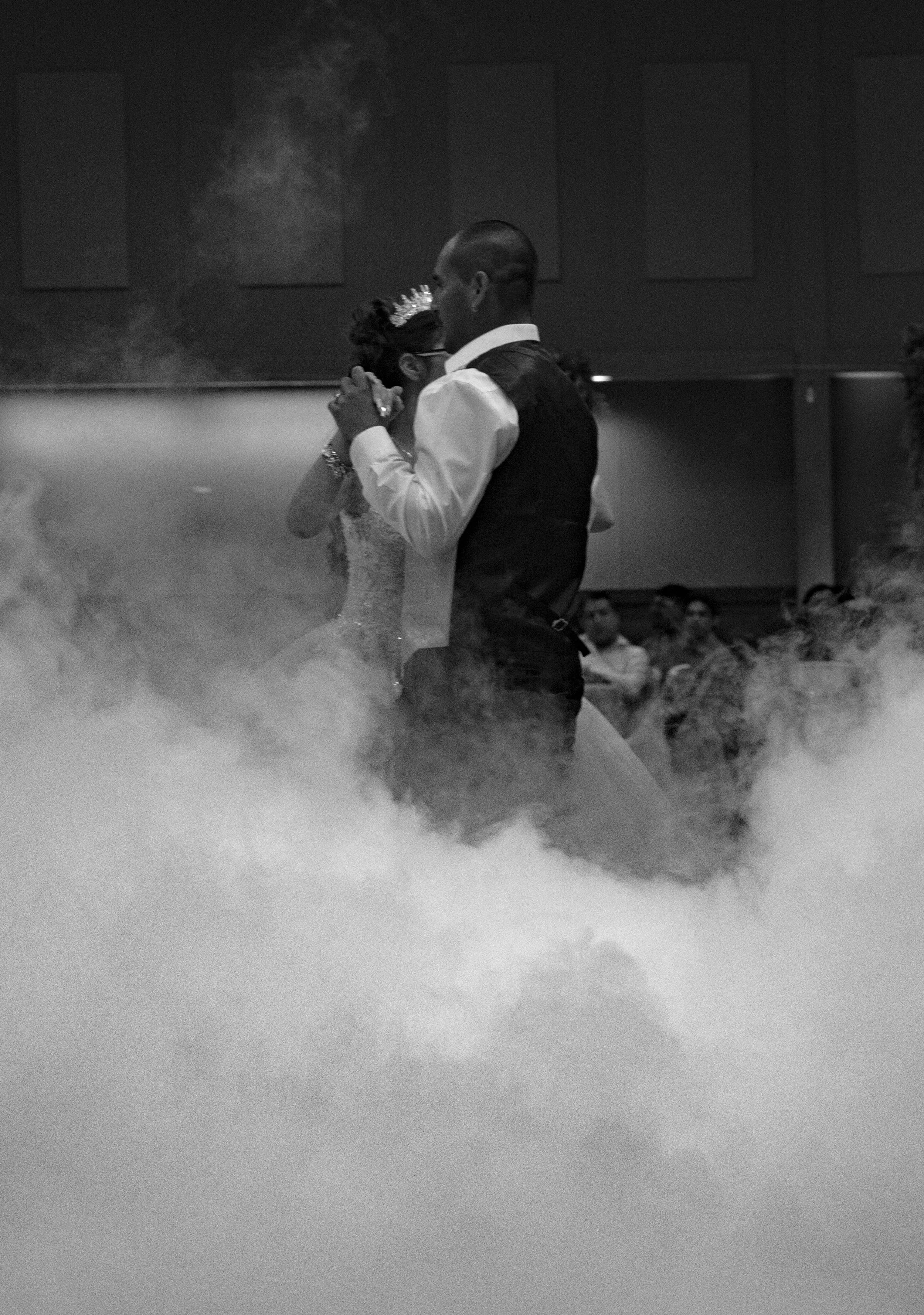 A couple dancing at a wedding reception, surrounded by fog and smoke, with guests watching in the background.