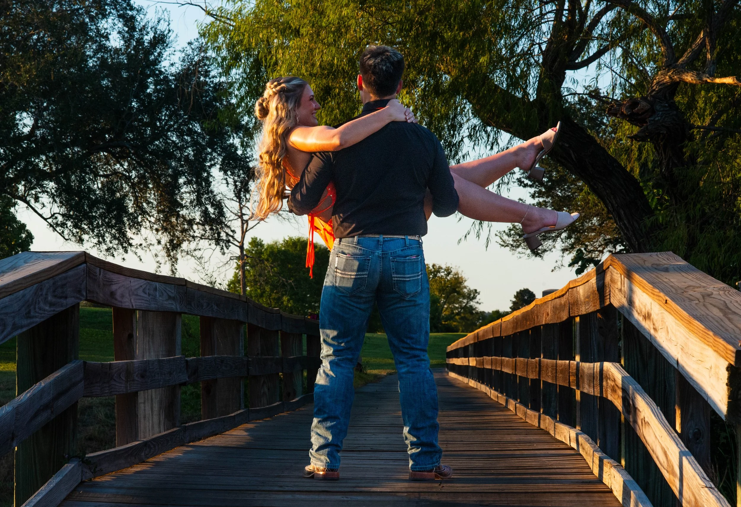 A young couple, man with dark hair in jeans and a woman with long curly hair in a red dress, standing on a wooden bridge outdoors during sunset. The man is holding the woman in his arms, and they are looking at each other.