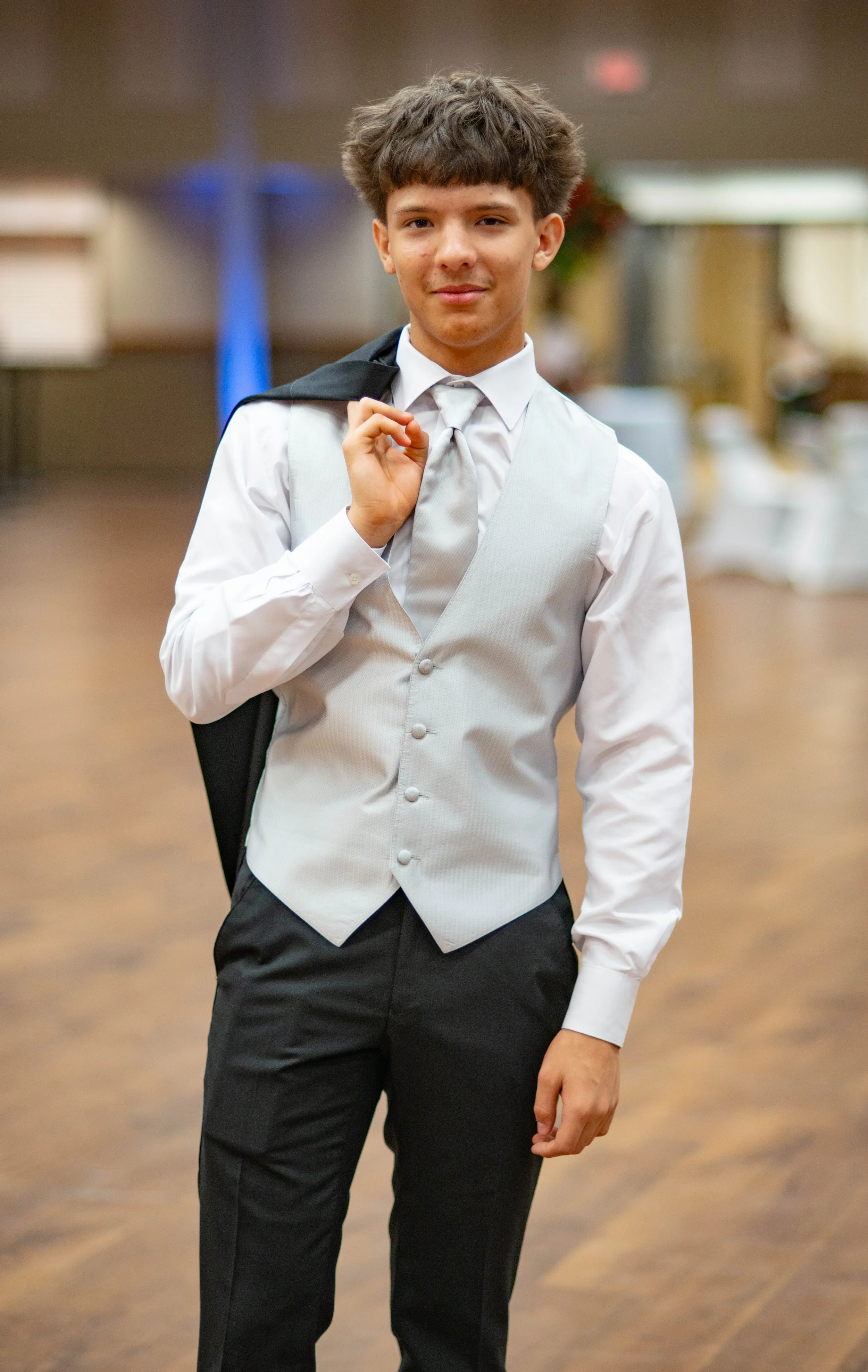 A young man dressed in formal attire, holding a black suit jacket over his shoulder, standing in an indoor setting with blurred background.